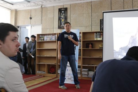 A man in a blue t-shirt with 'Startup' written on it speaks into a microphone in front of a group of people.