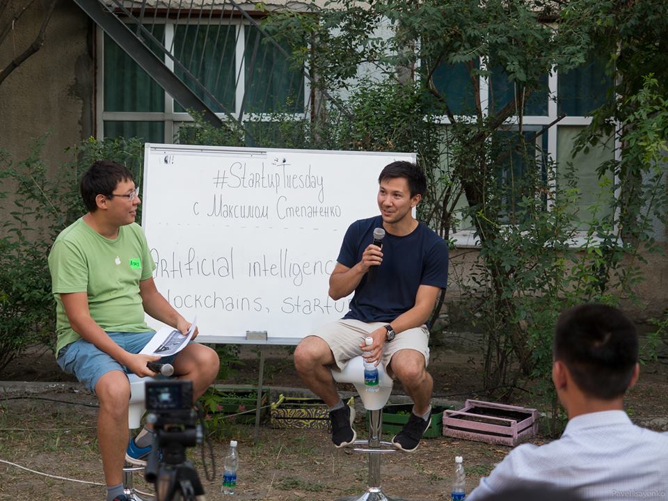 Two men sit on stools in front of a whiteboard discussing AI, blockchain, and startups.