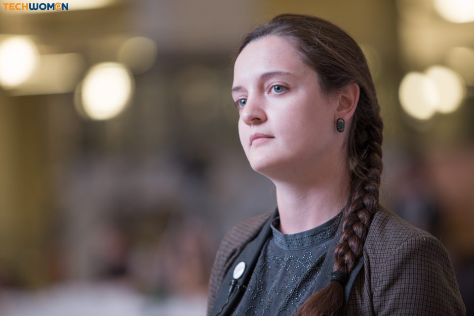 A woman with a braided hairstyle and a button on her blazer, in a setting with blurred lights.