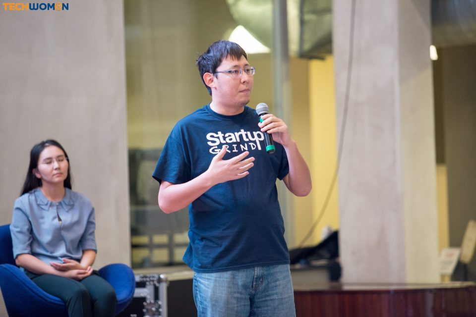 A man in a 'Startup Grind' t-shirt speaks into a microphone at an event.
