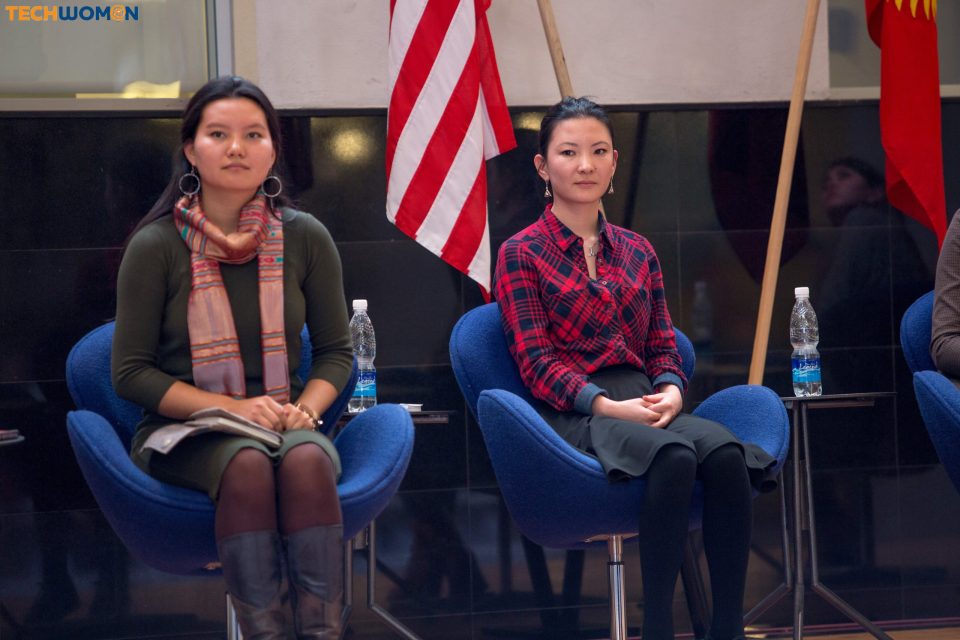 Two women seated in chairs in front of flags, possibly at a conference or event.