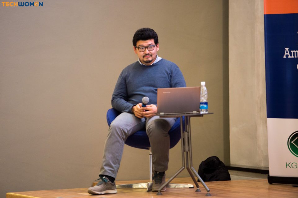 A person sitting on a stage with a laptop and a microphone, likely participating in a tech-related event.