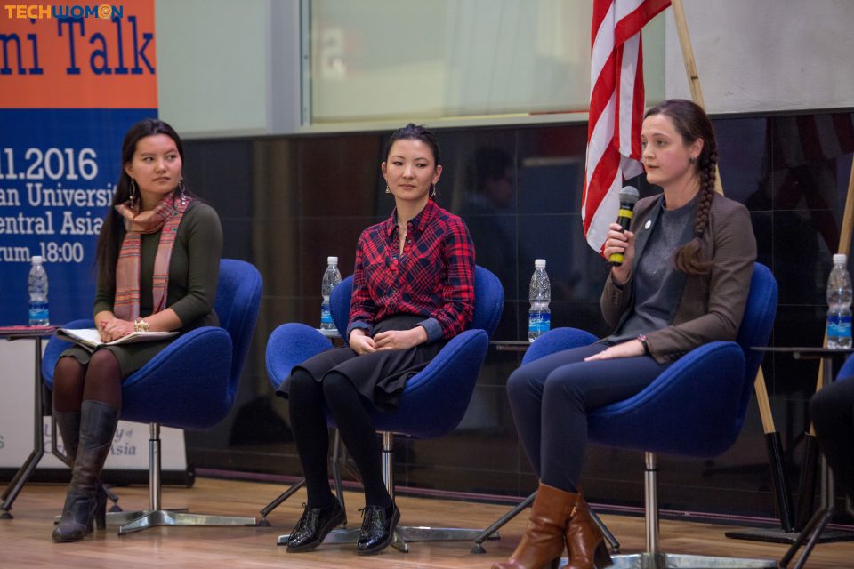 A woman stands at a microphone in a conference room with chairs and a banner in Kyrgyzstan.