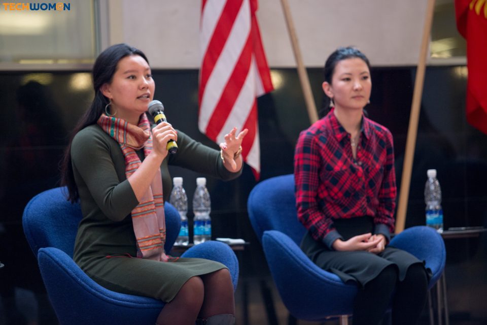 Two women seated in blue chairs, one speaking into a microphone with a flag in the background.