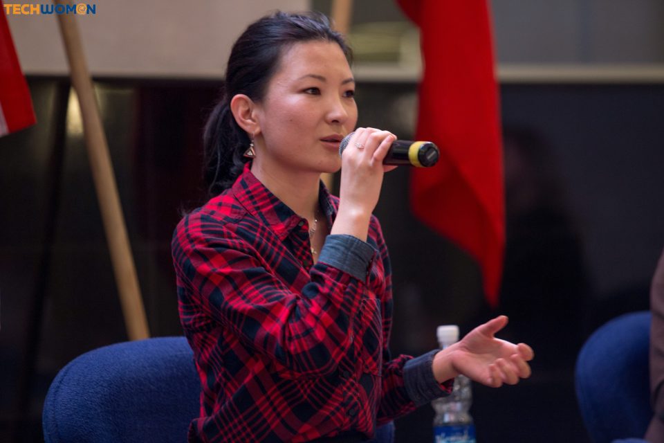 A woman in a red and black plaid shirt speaks into a microphone during a panel discussion.