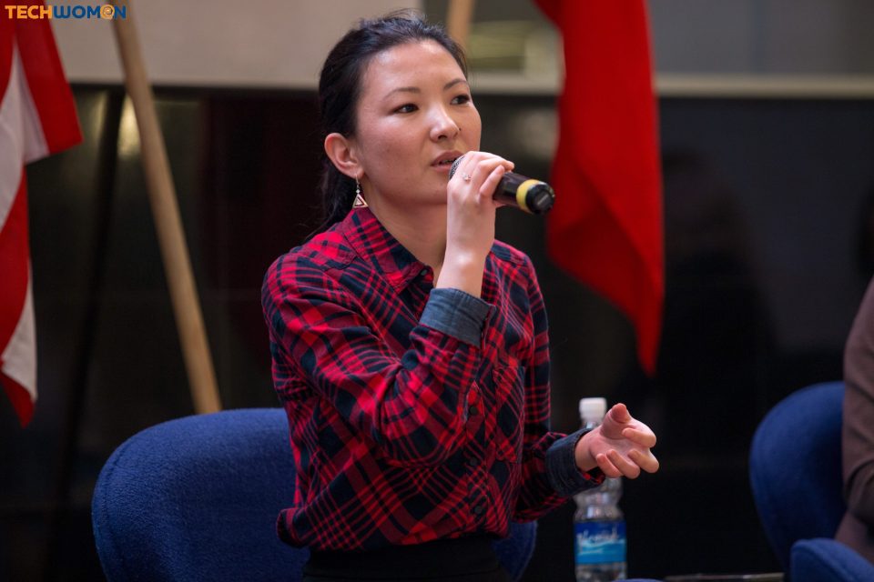 A woman in a red and black plaid shirt speaks into a microphone at an event in Bishkek, Kyrgyzstan.