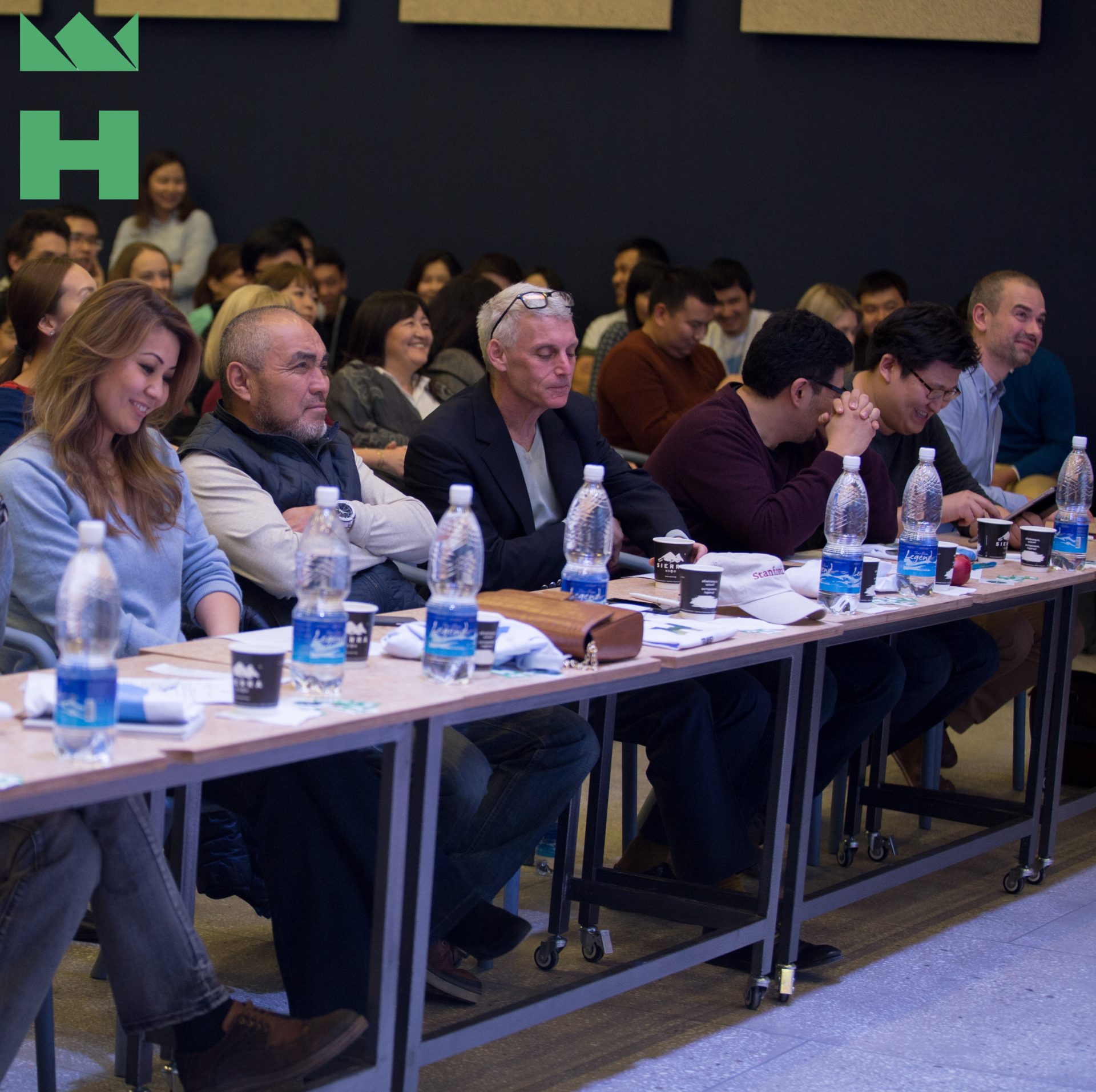 Participants seated at tables in a conference setting, with water bottles and cups.