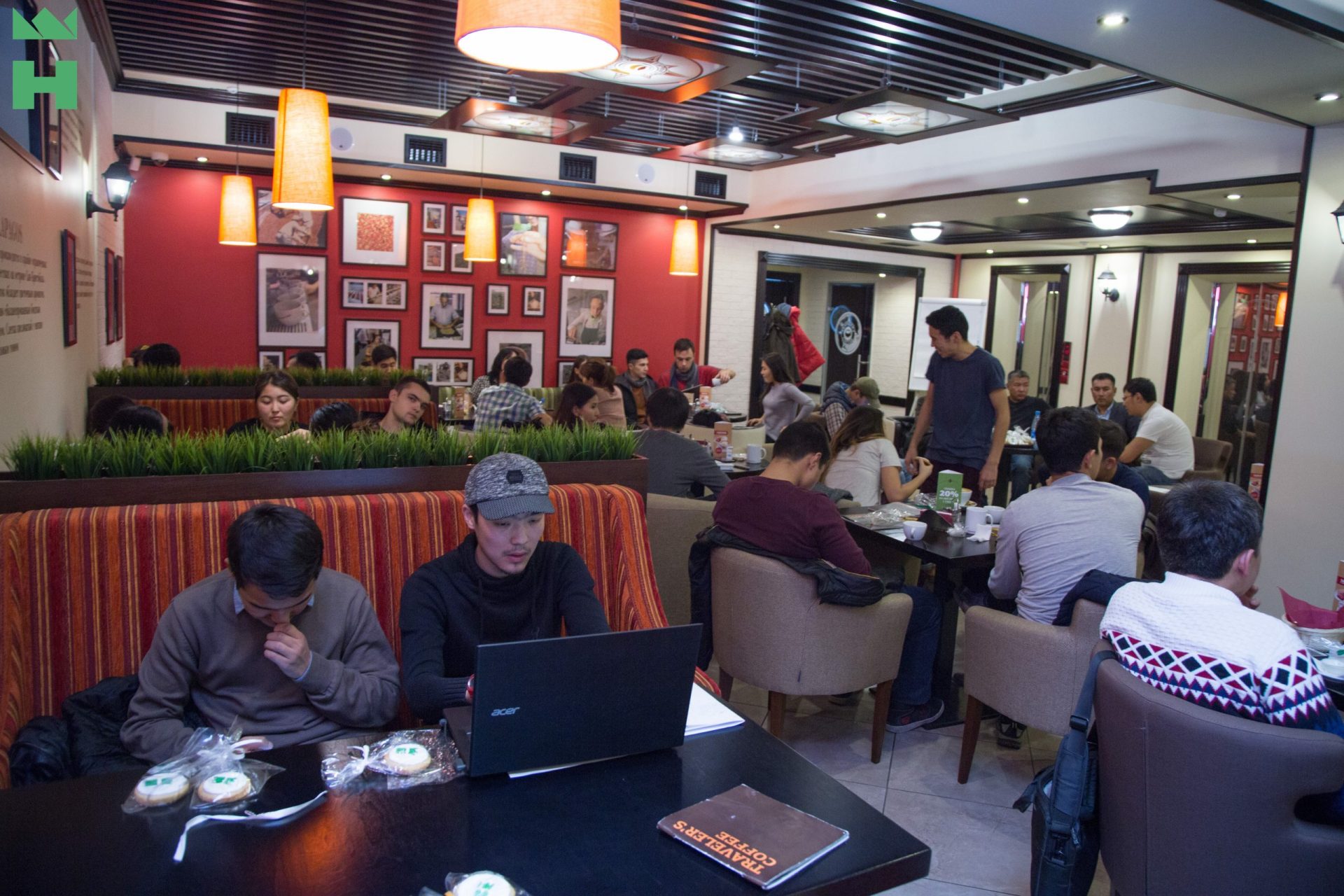 A group of people sitting at tables in a cozy café, engaged in various activities such as working on laptops and enjoying food and drinks.