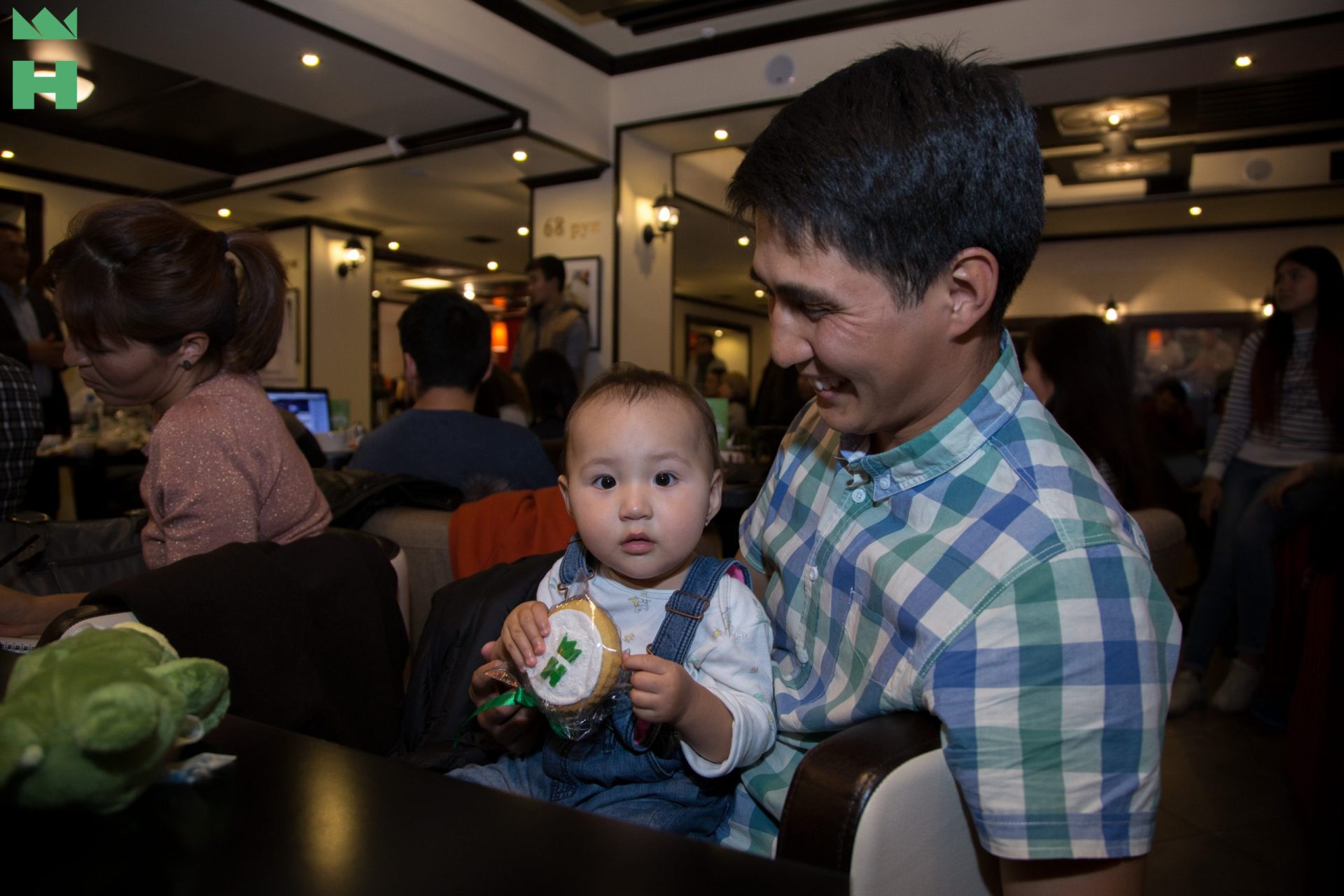 A man and a child sit together at a table in a restaurant, with the child holding a wrapped cookie.