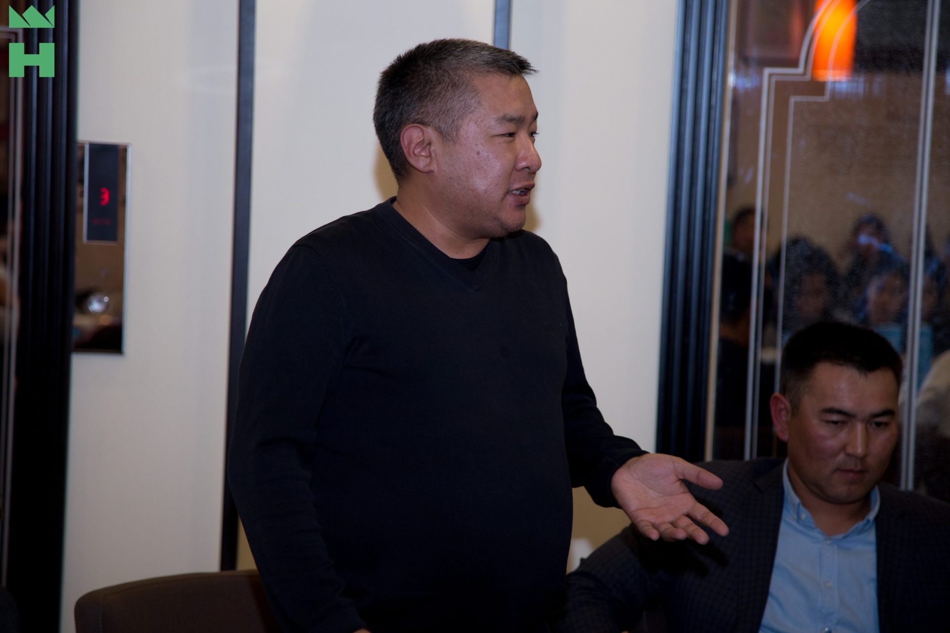 A man in a black sweater gestures while standing in a room with another person seated in the background.