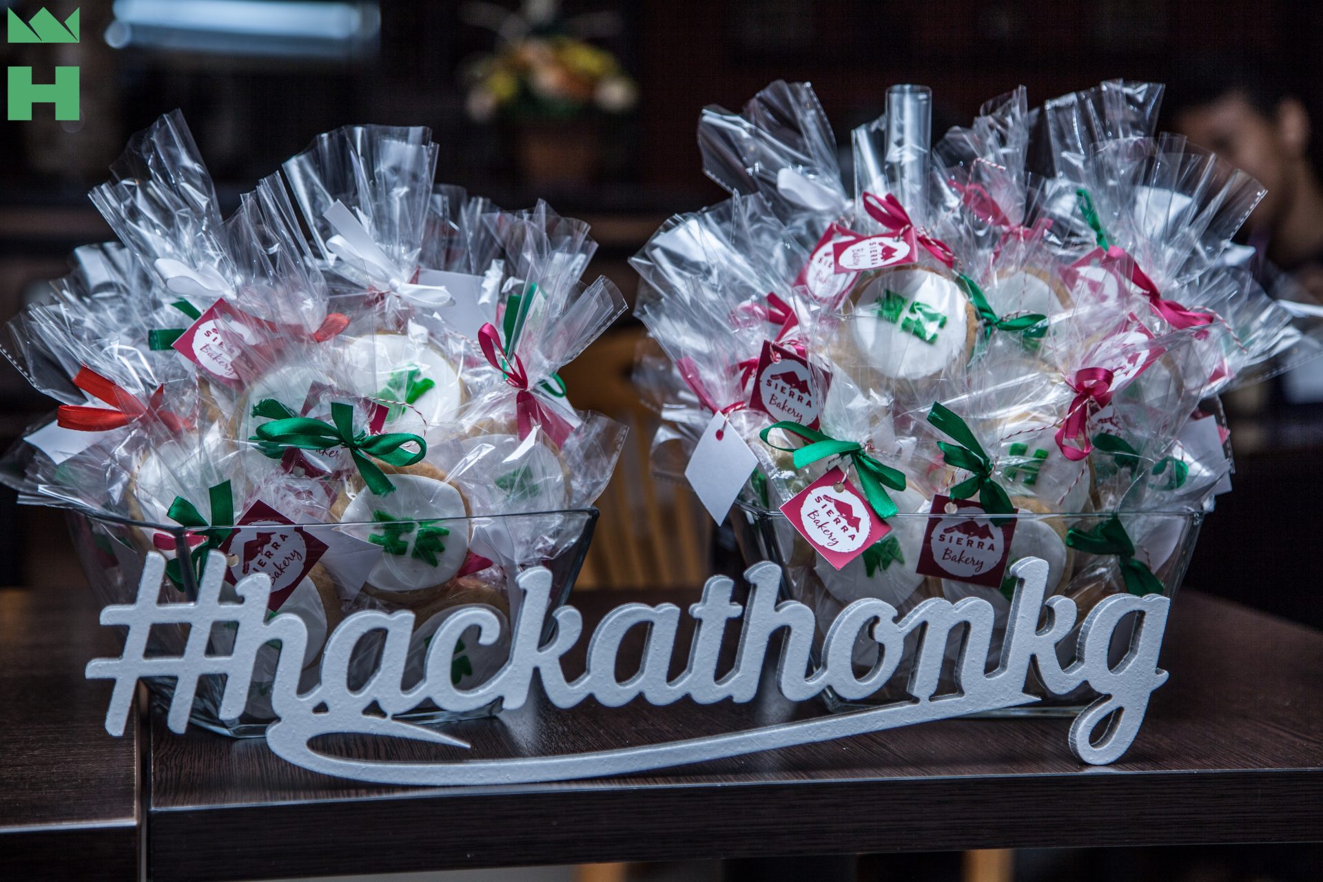 Bakery items in clear plastic bags with ribbons and tags, displayed on a table with a banner reading '#hackathonkg'.