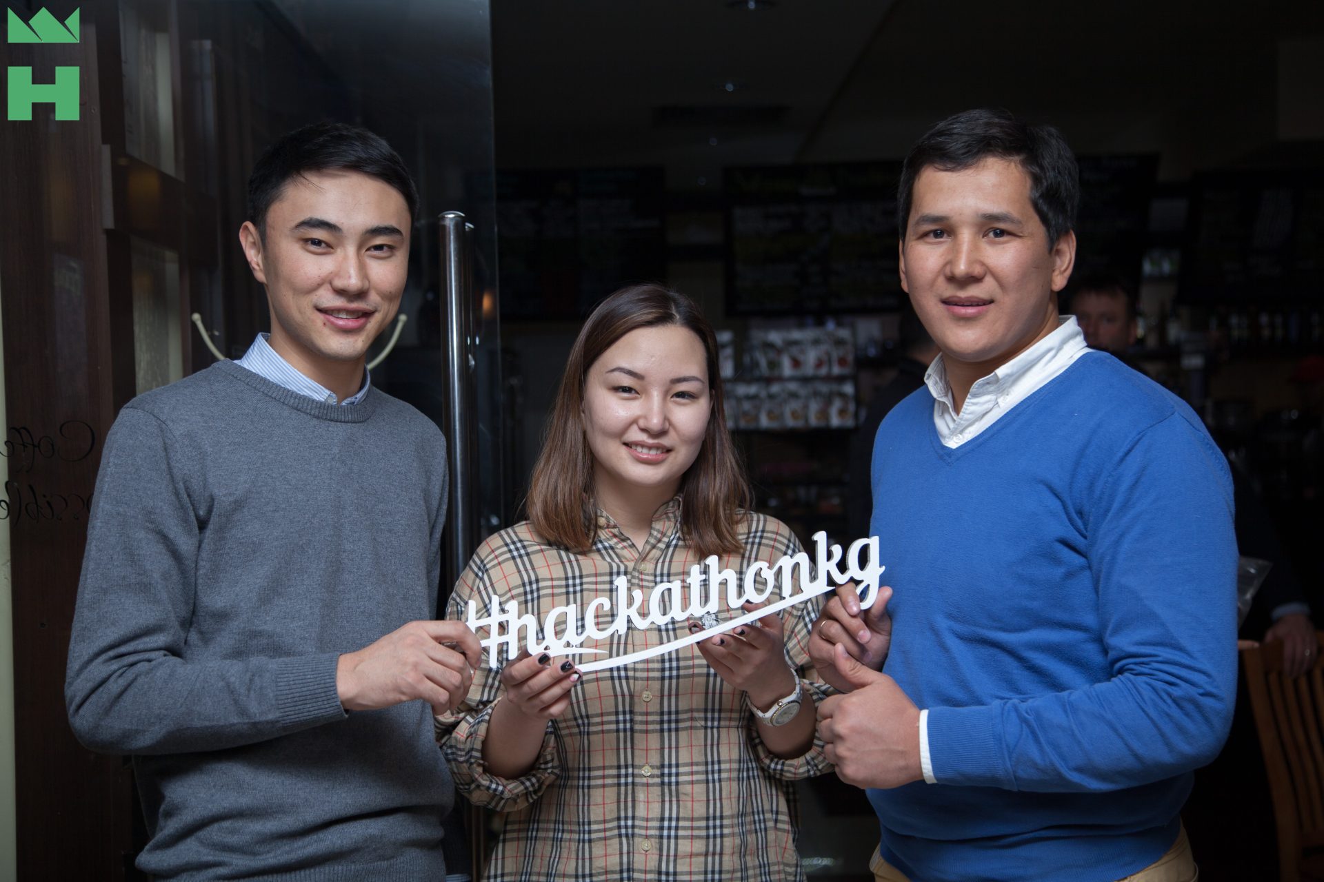 Three individuals holding a sign that reads '#hackathonkg' at an event in Bishkek, Kyrgyzstan.