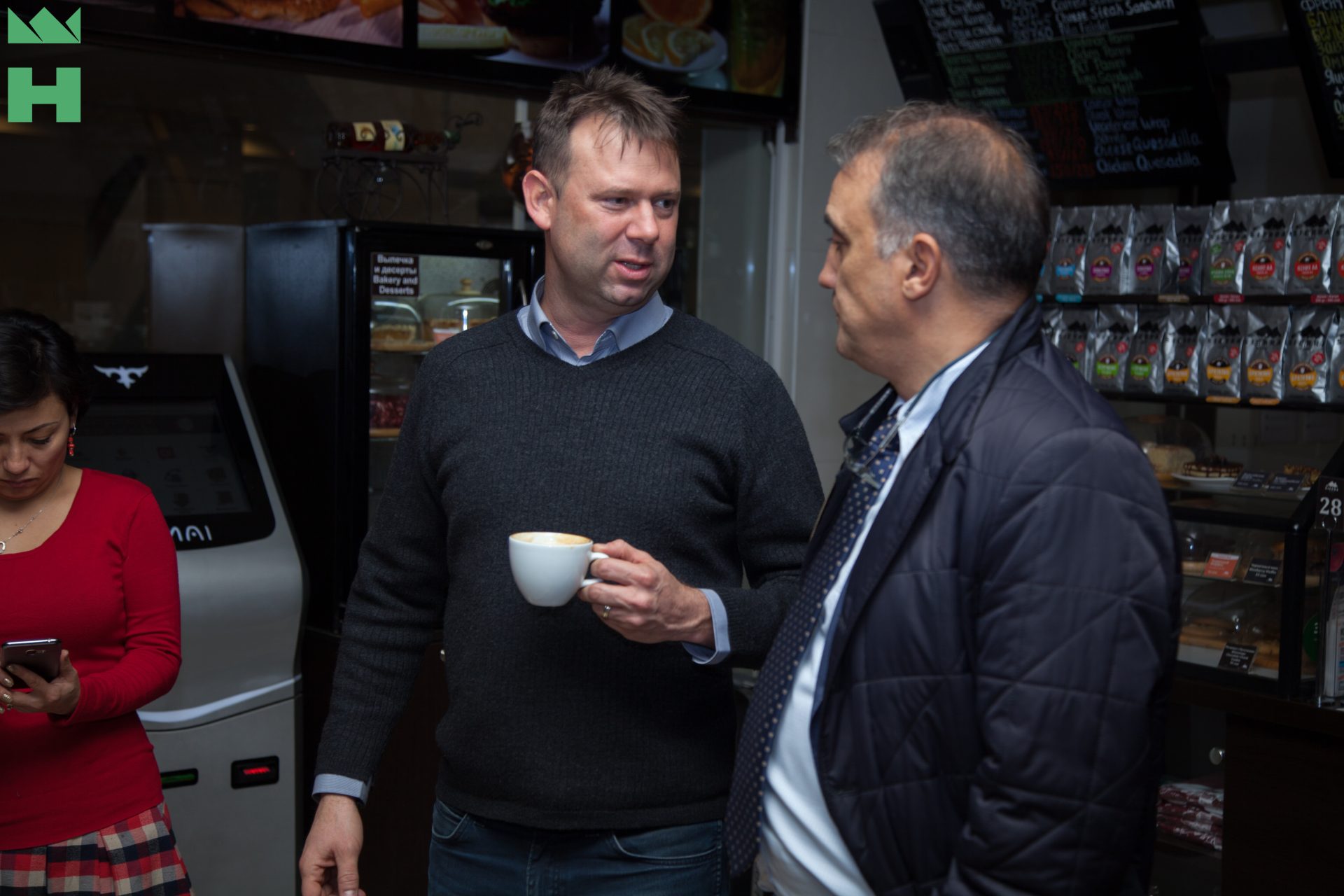 Two men having a conversation in a cafe, one holding a coffee cup.