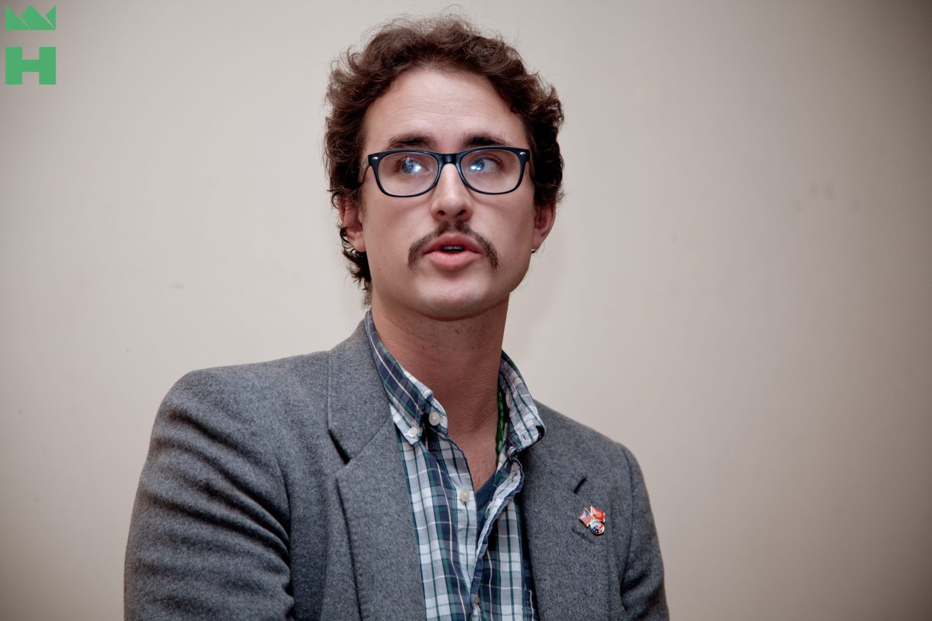 A person with a mustache and glasses is wearing a gray blazer over a plaid shirt.