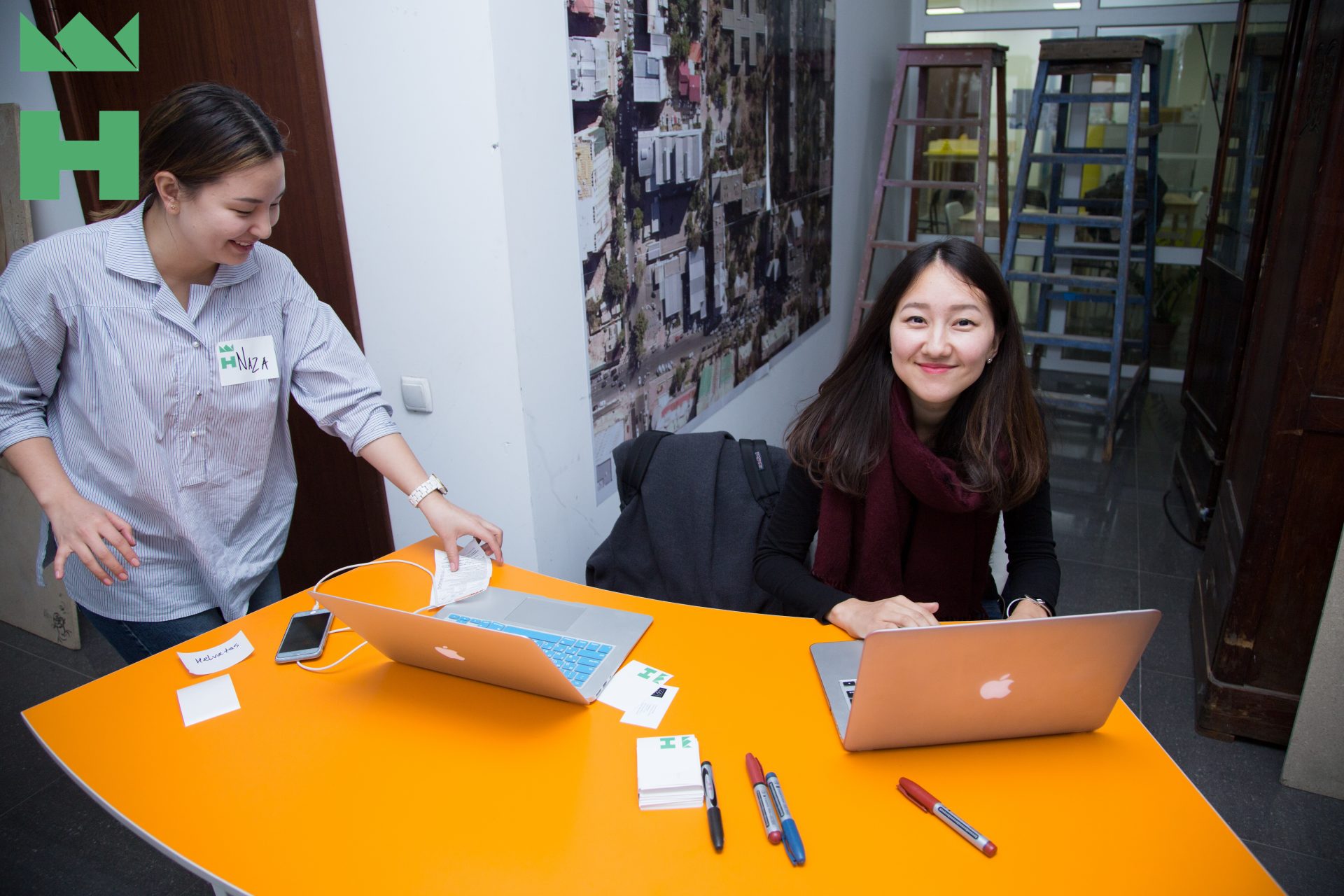 Two individuals working on laptops at an orange table in a workshop setting.