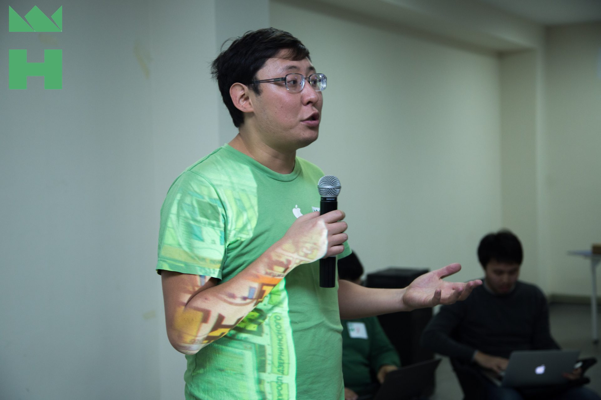A person speaking at an event in Bishkek, Kyrgyzstan, holding a microphone and wearing a green t-shirt.