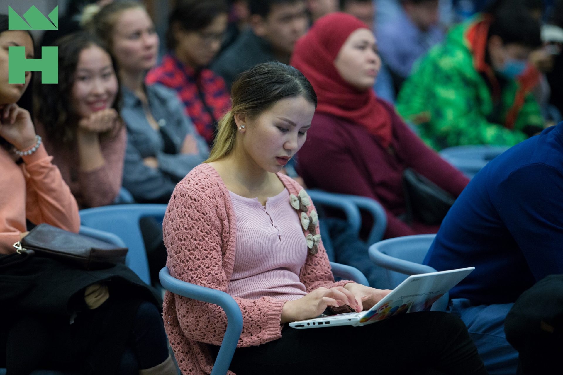 A young woman in a pink sweater uses a laptop in a crowded auditorium.