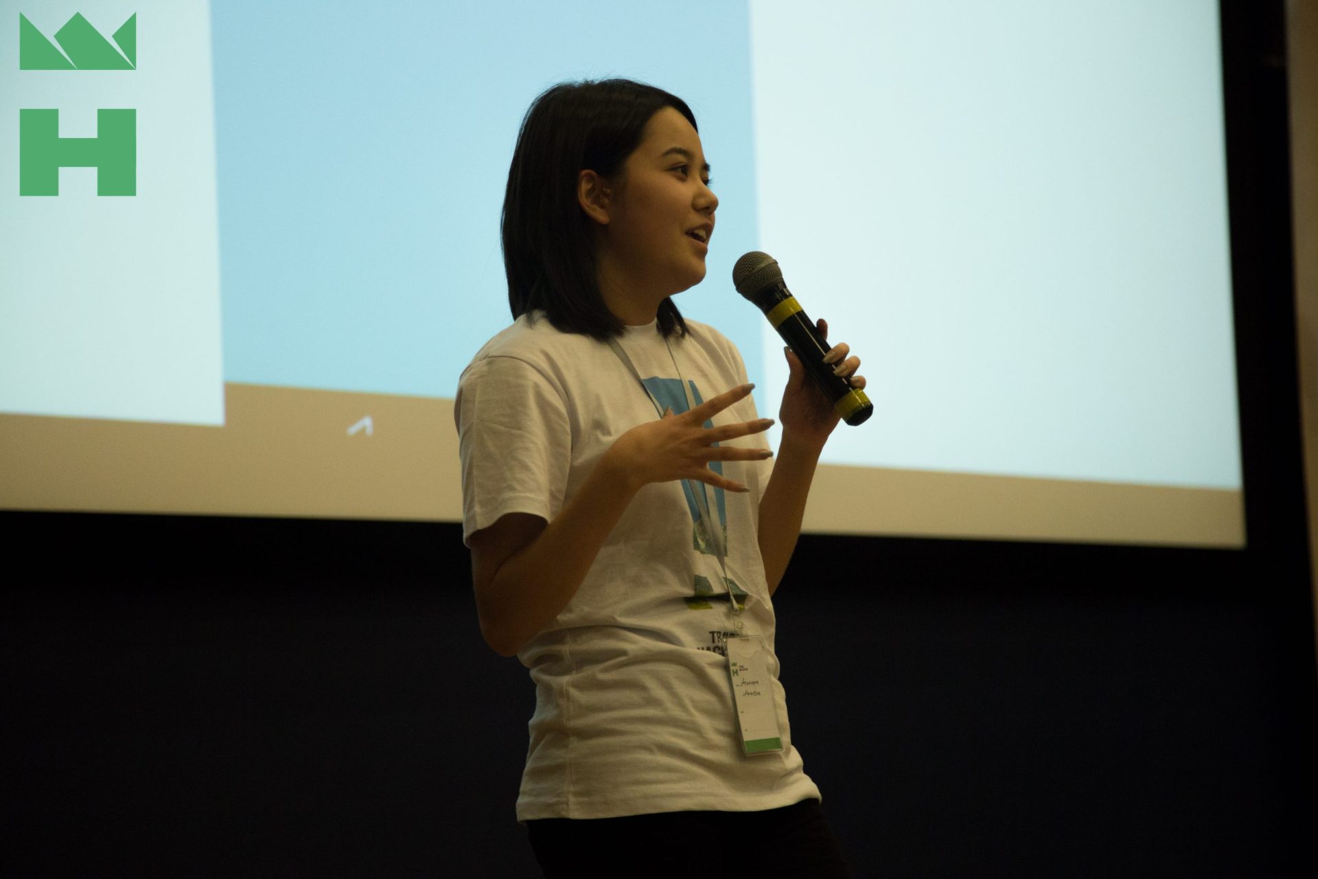 A young woman speaks into a microphone on stage with a projector screen in the background.