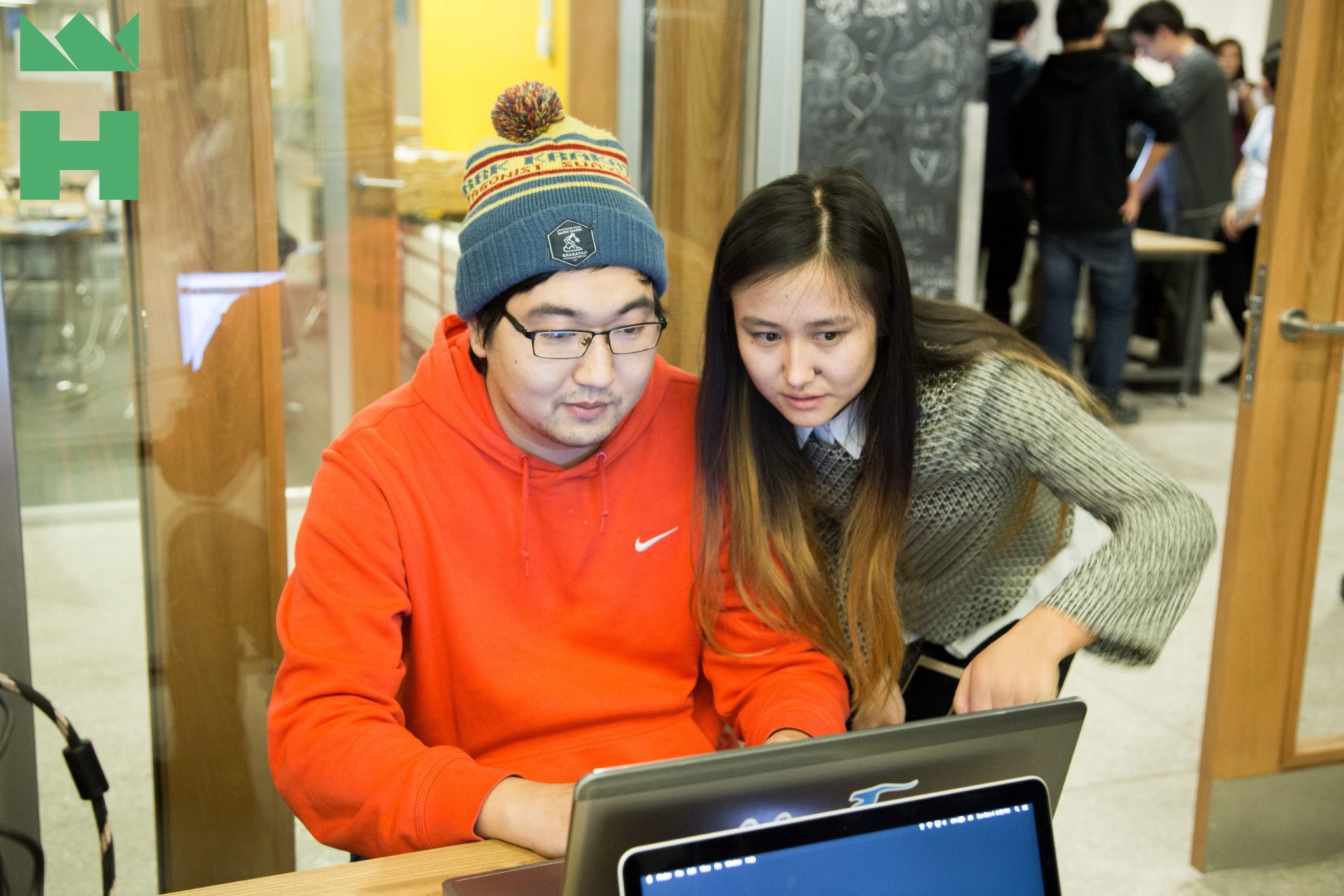 Two individuals working together on a laptop in a busy office setting.