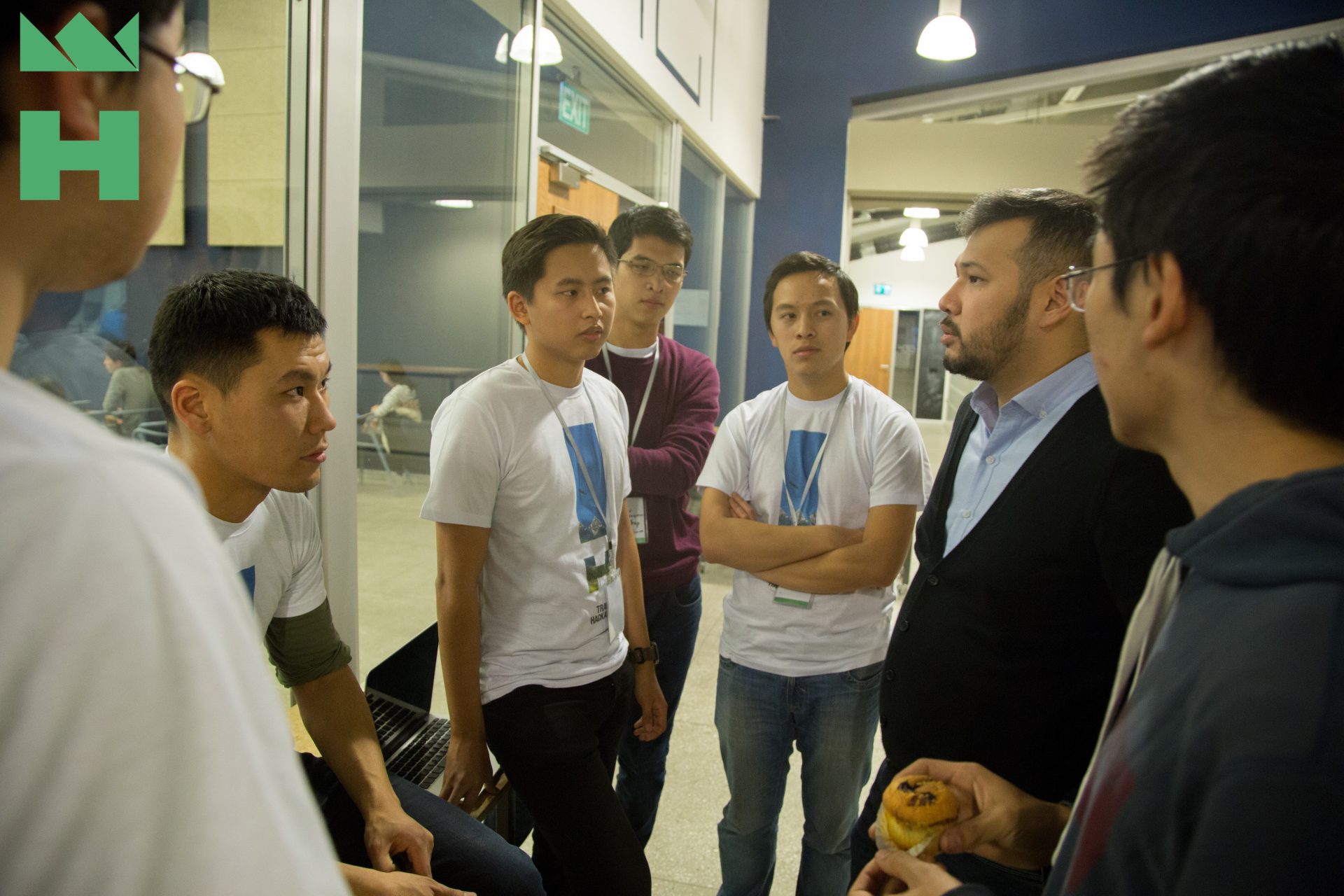 A group of people in white t-shirts with a logo on them are gathered in a room, possibly discussing a project or idea.