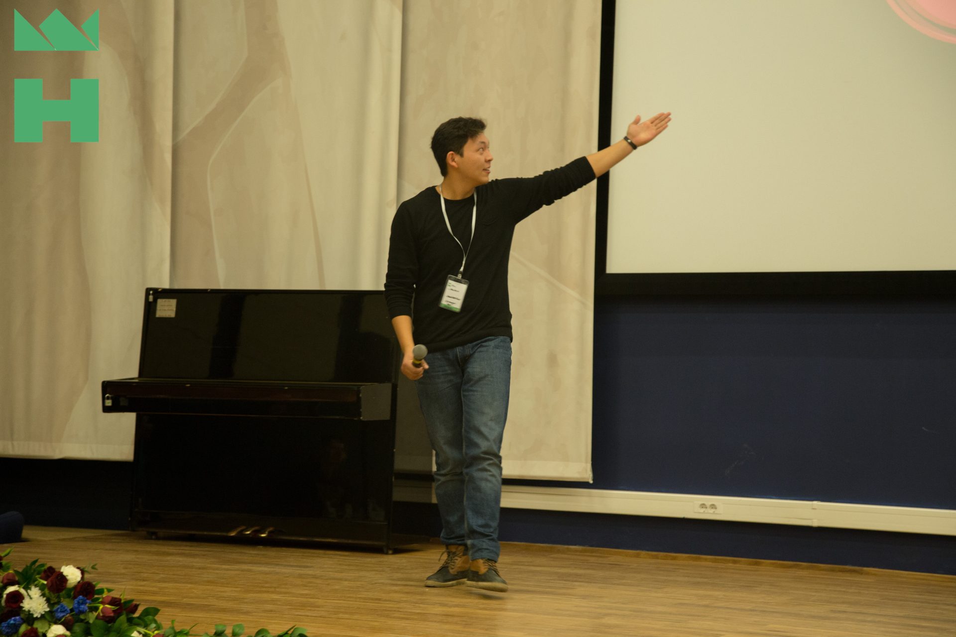 A person giving a presentation on stage with a piano and a projector screen in the background.