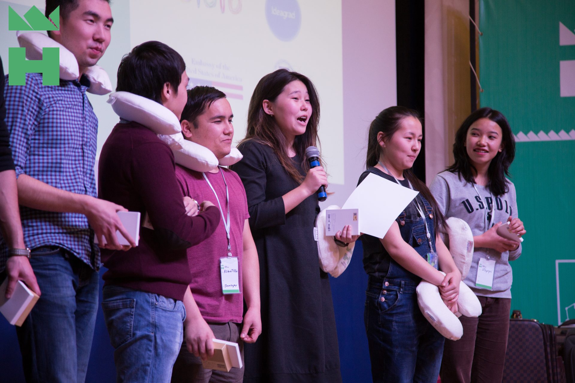 A group of young people stands on stage, holding pillows and boxes, with one person speaking into a microphone.