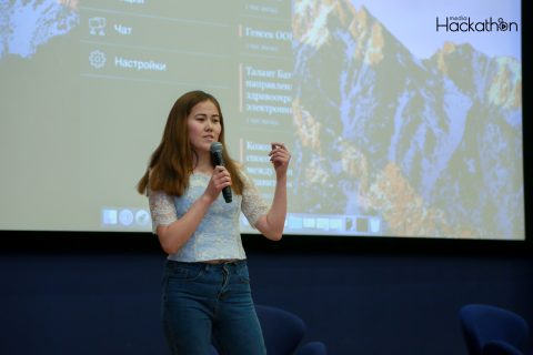 A person speaking into a microphone on stage with a presentation screen displaying text and a mountain image in the background.