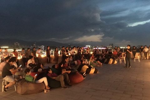 A large group of people sitting on bean bags and chairs on a rooftop at night, with a city skyline in the background.