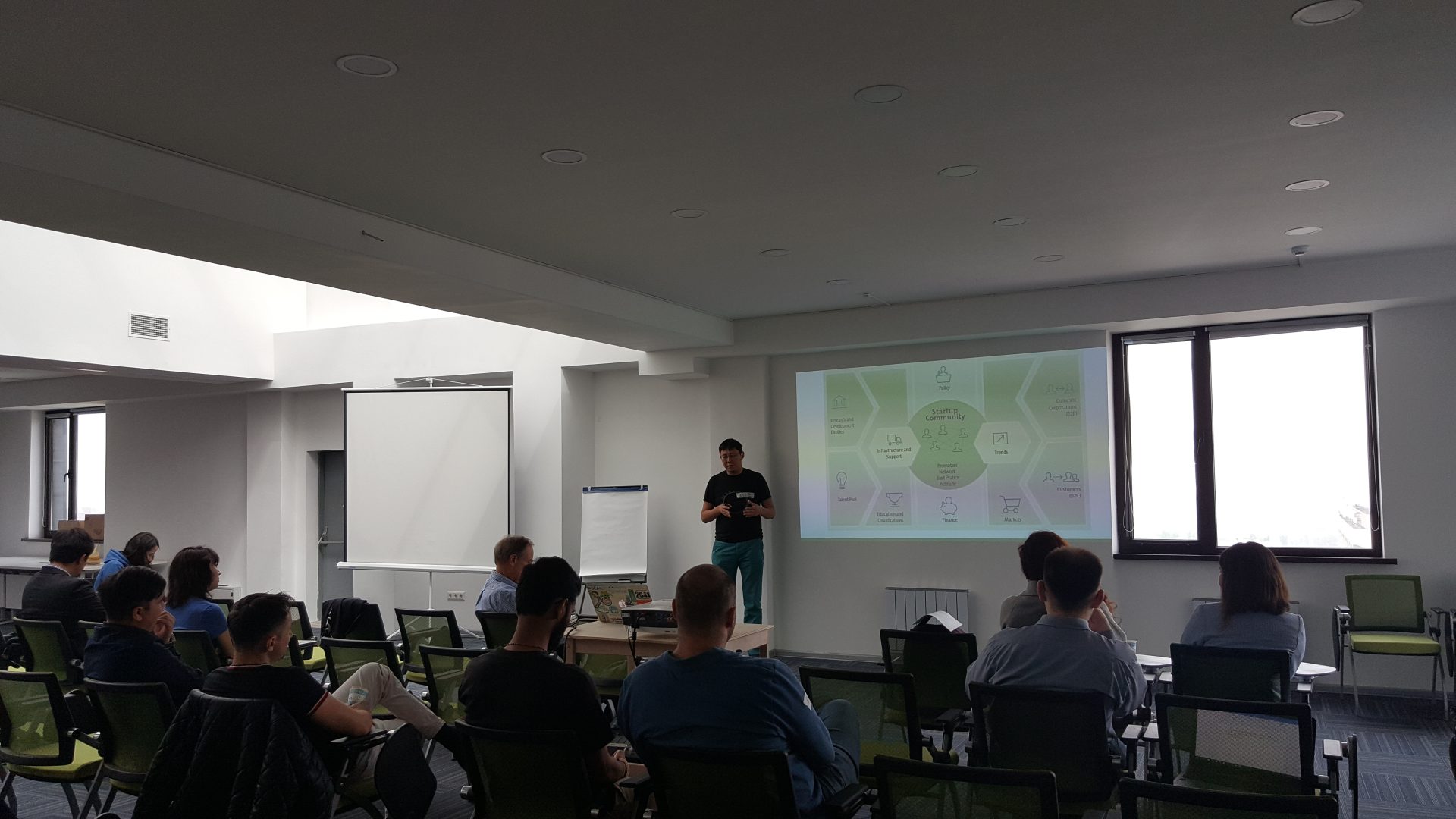A workshop or conference is taking place in a modern room with participants seated and a presenter standing in front of a screen displaying a diagram.
