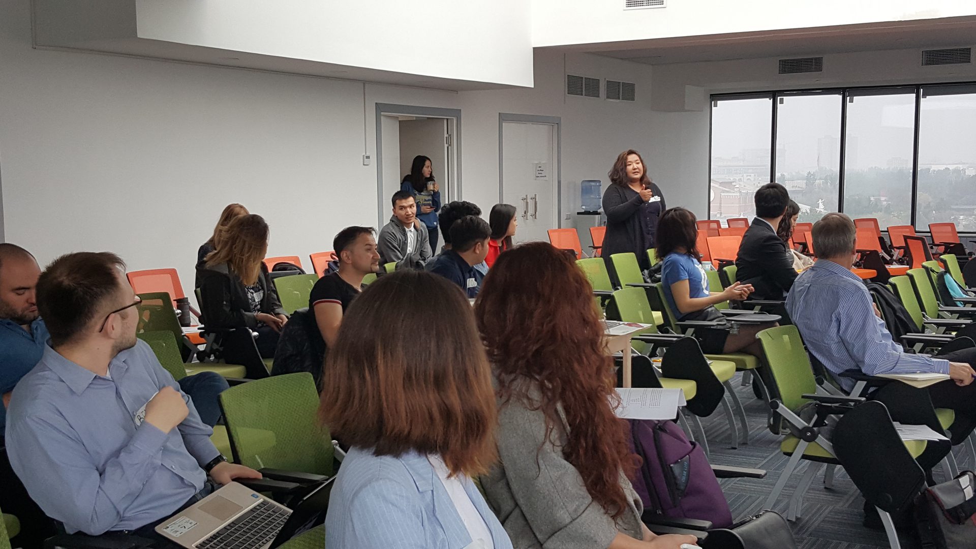 Participants in a workshop or conference in a modern, well-lit room with green chairs and large windows.
