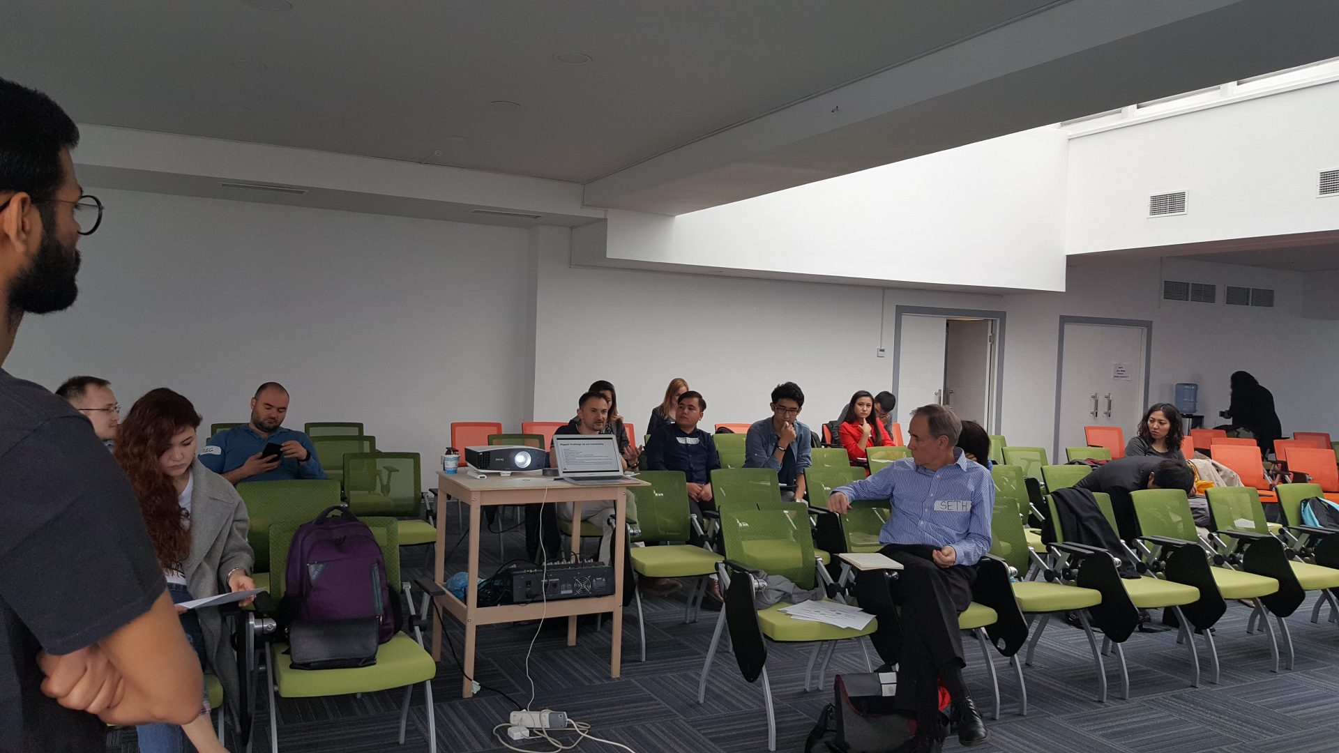 Participants in a workshop or conference in a modern, well-lit room with green chairs and a projector.