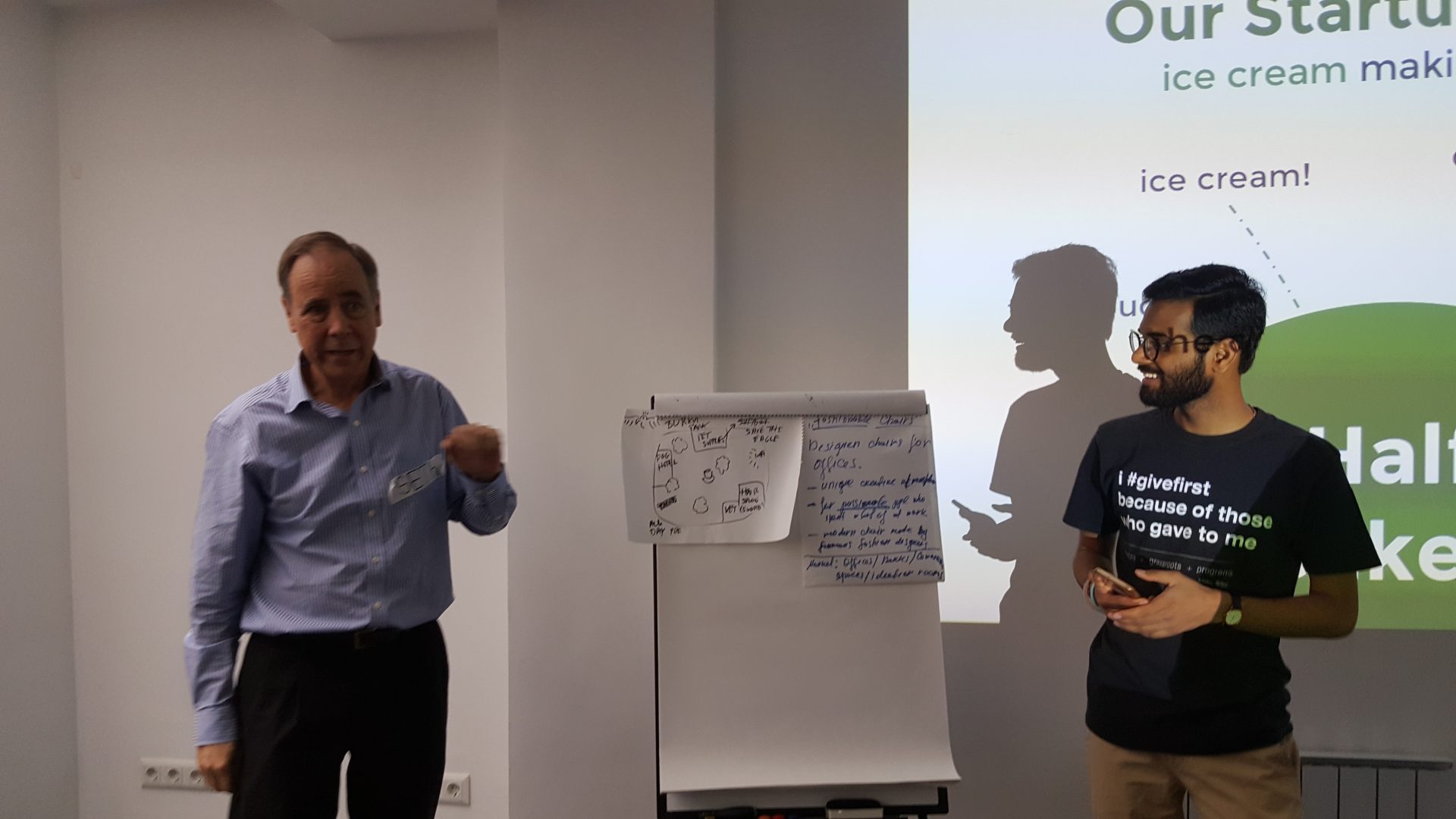 Two individuals are presenting in a workshop setting with a flip chart and a projected slide.