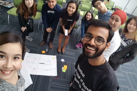 A group of people in a classroom setting, with a whiteboard and sticky notes on the floor.