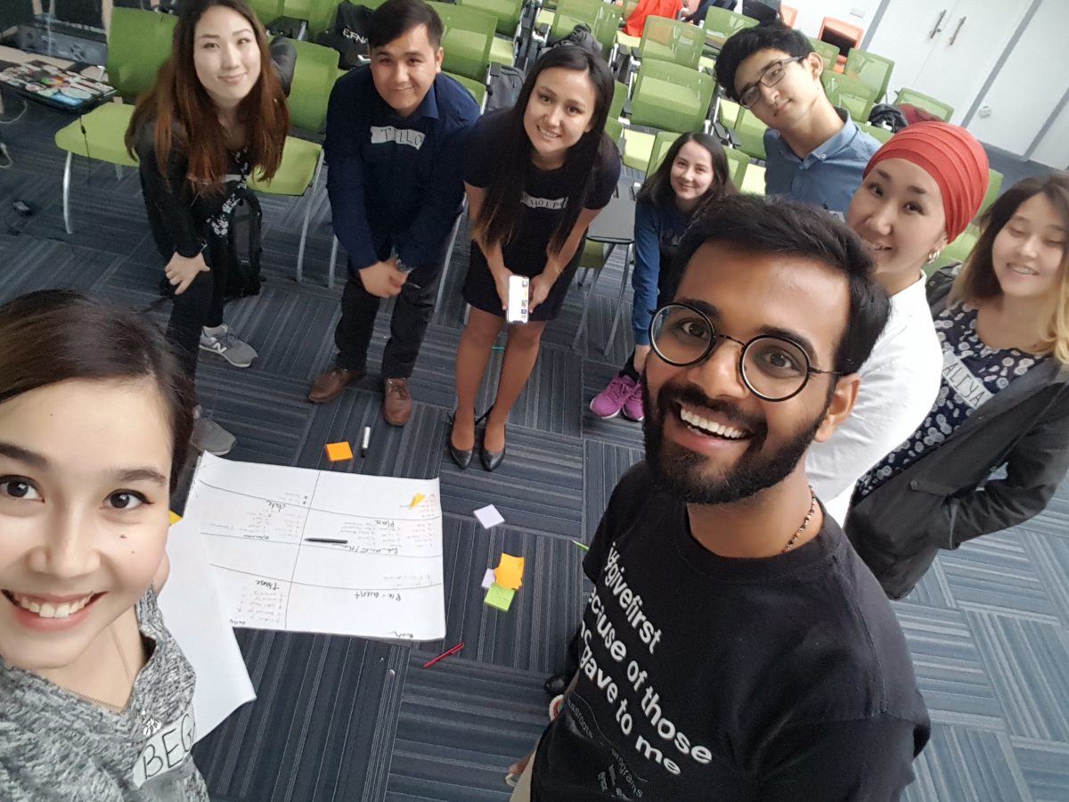A group of people in a classroom setting, with a whiteboard and sticky notes on the floor.