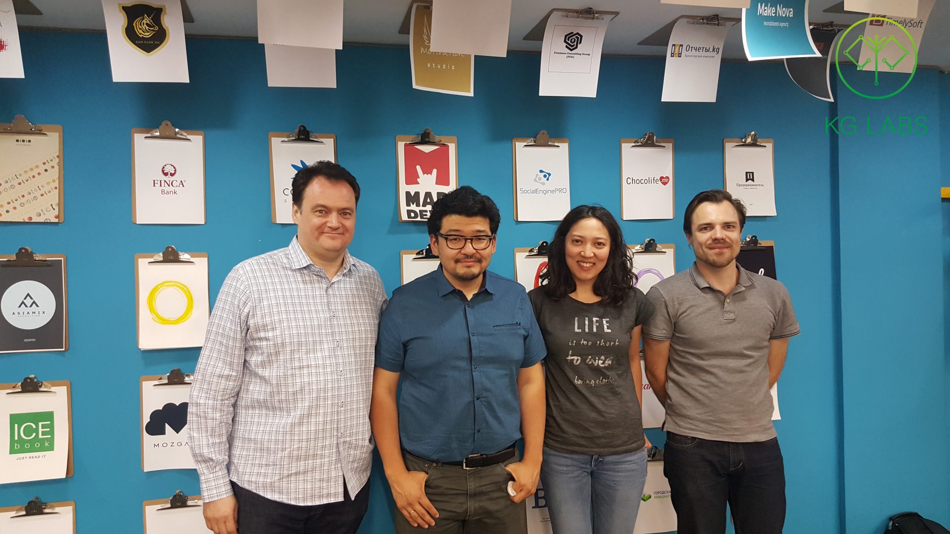 Four individuals posing for a photo in front of a blue wall with various logos and clipboards.