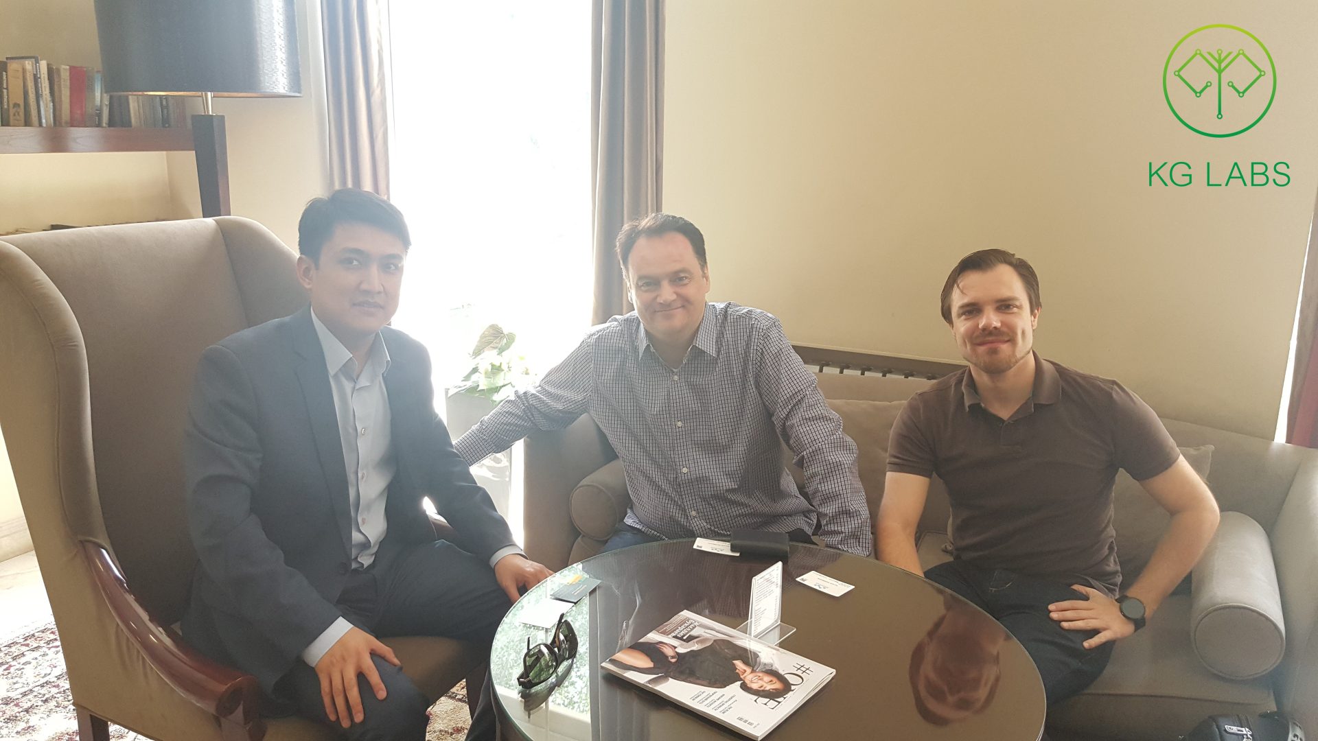 Three individuals sitting in a meeting room with a table and magazines.