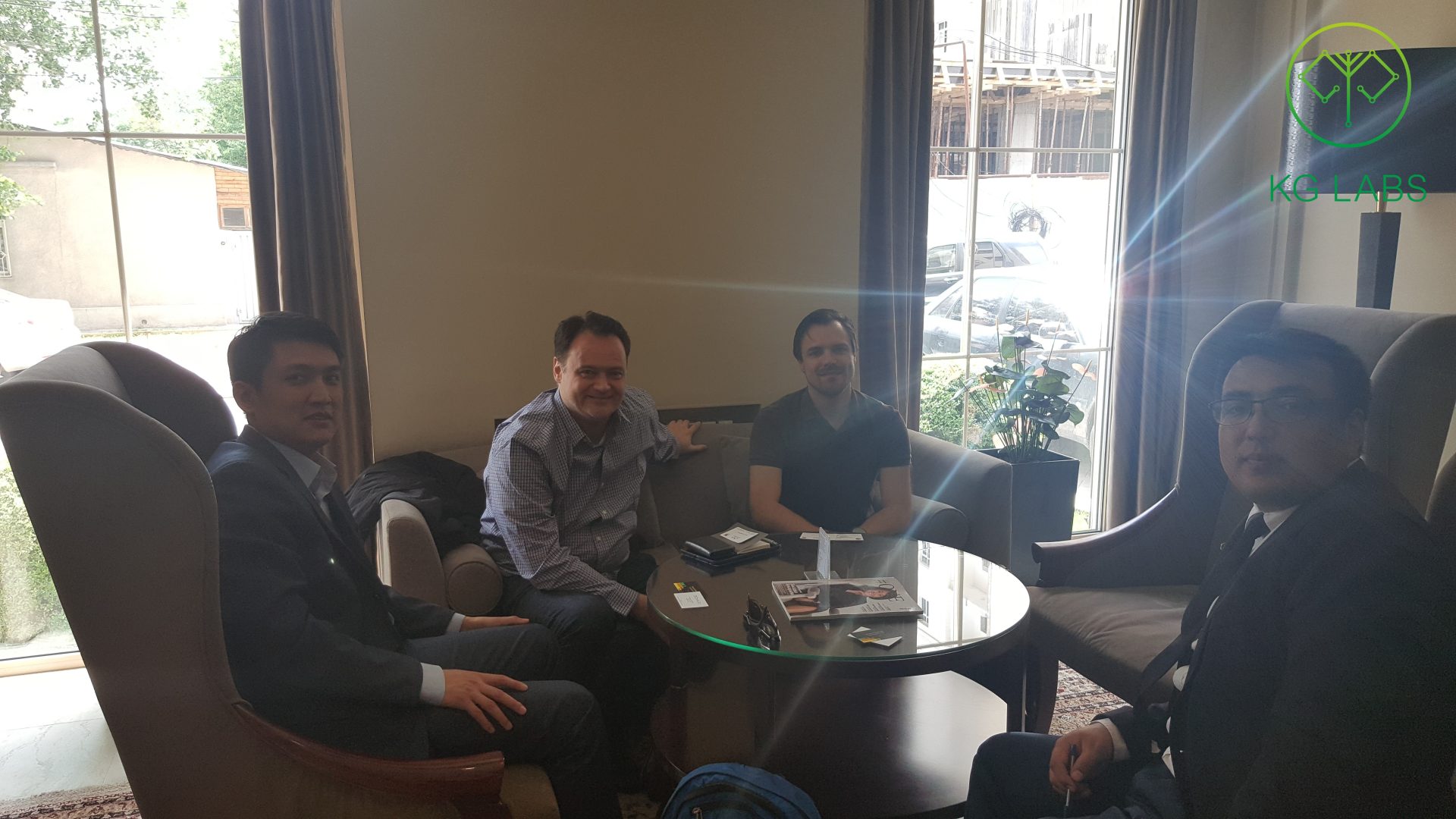Four individuals engaged in a meeting in a modern, well-lit room with a glass table and comfortable seating.