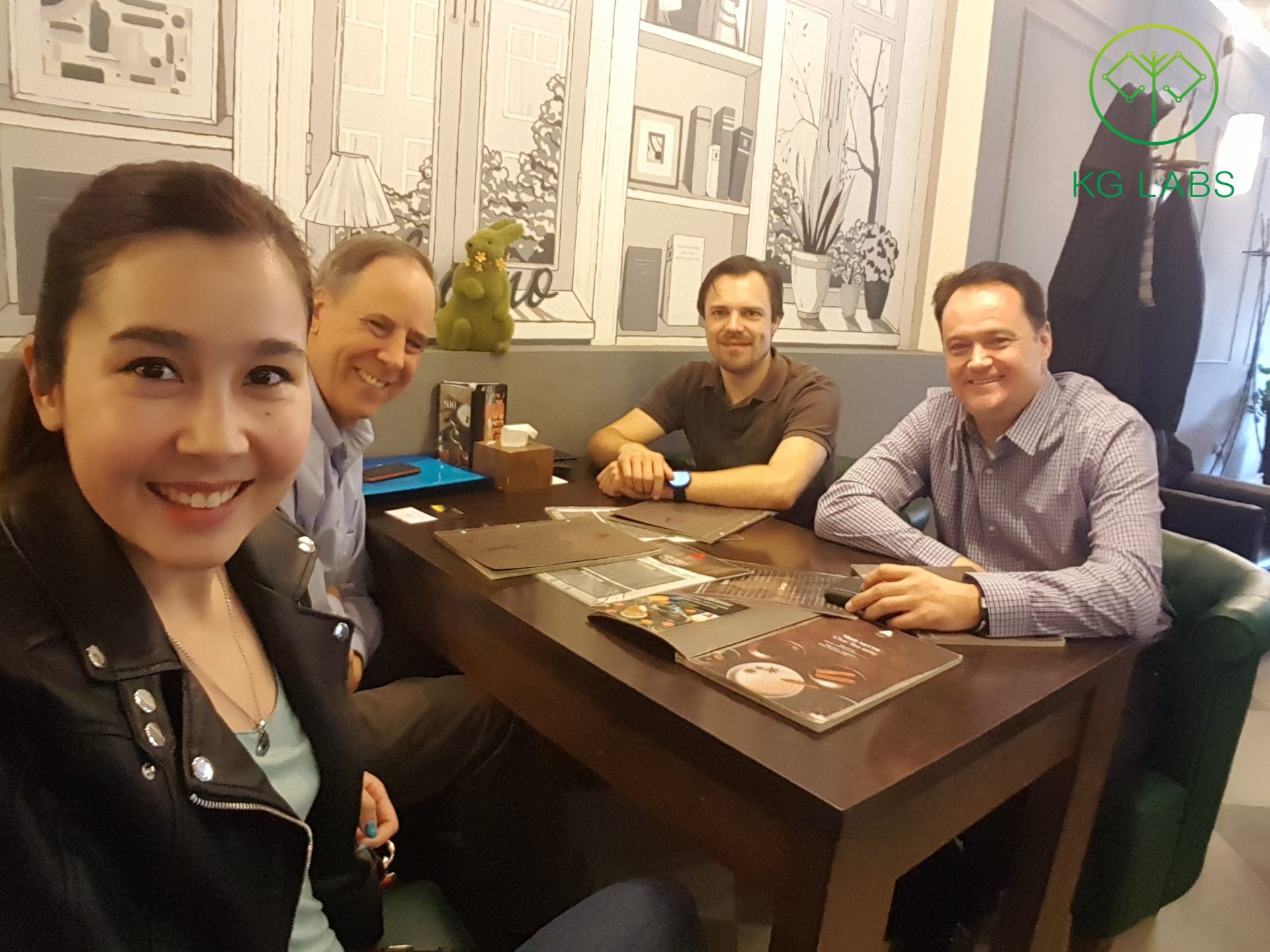 A group of four people sitting at a table in a cafe, smiling for a selfie.