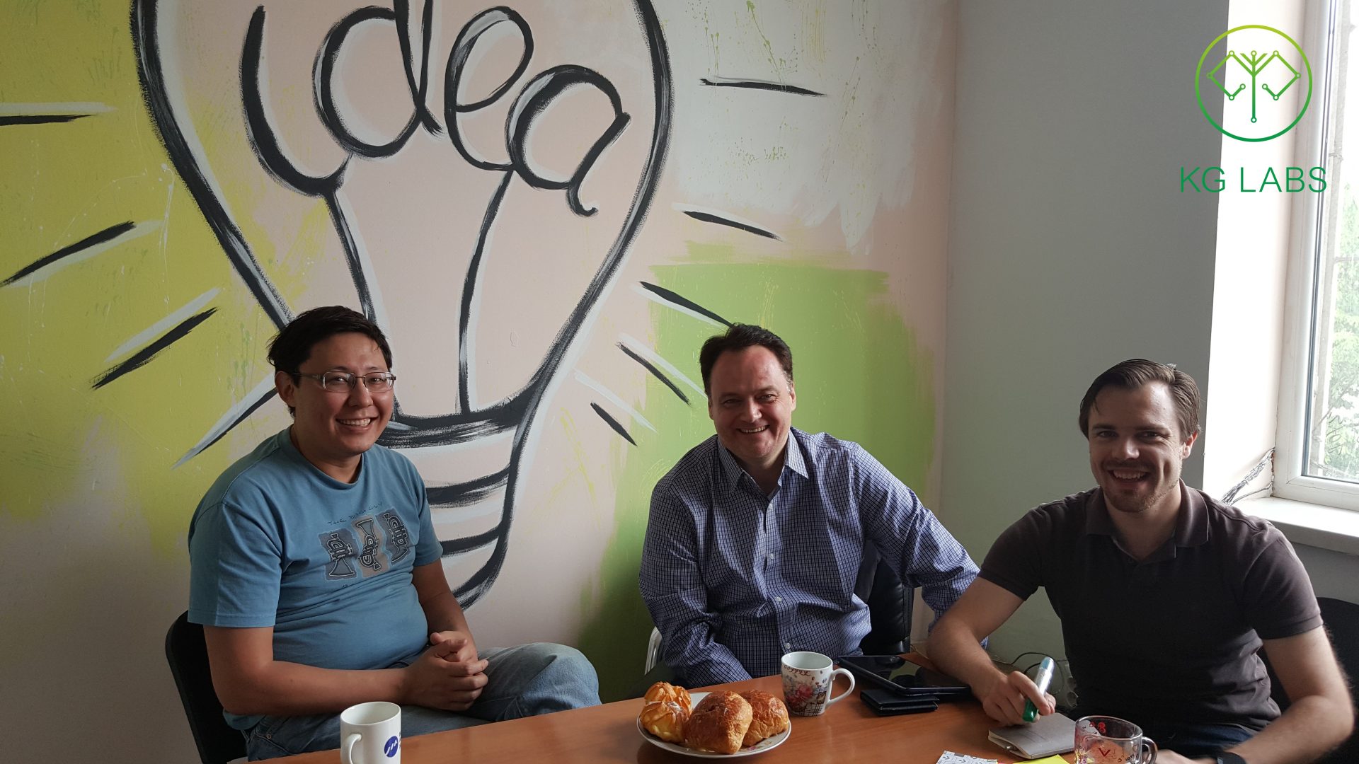 Three individuals sitting at a table in a room with a mural of a light bulb and the word 'idea' on the wall.
