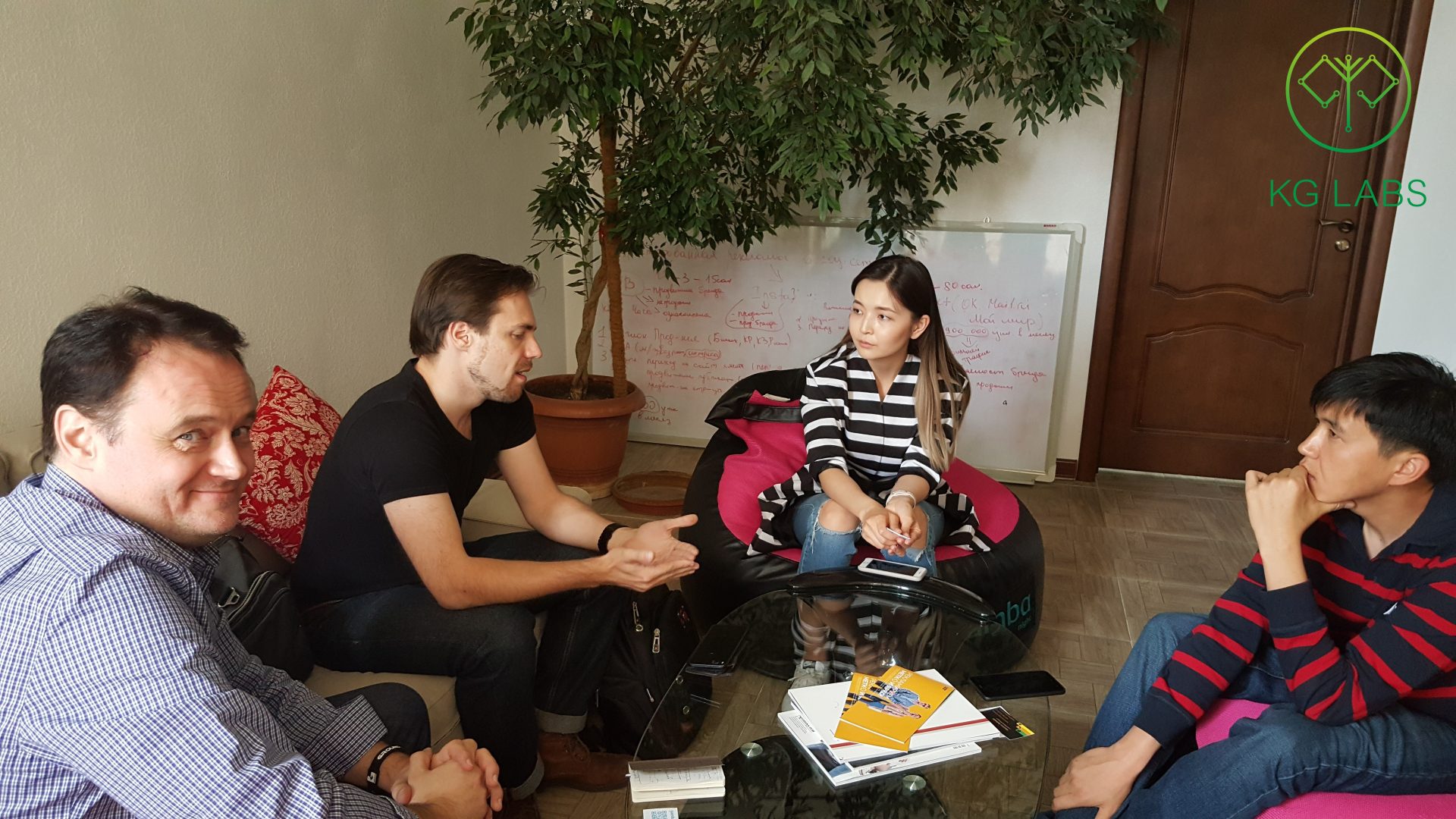 Four individuals engaged in a discussion in a relaxed setting with a whiteboard and potted plant in the background.