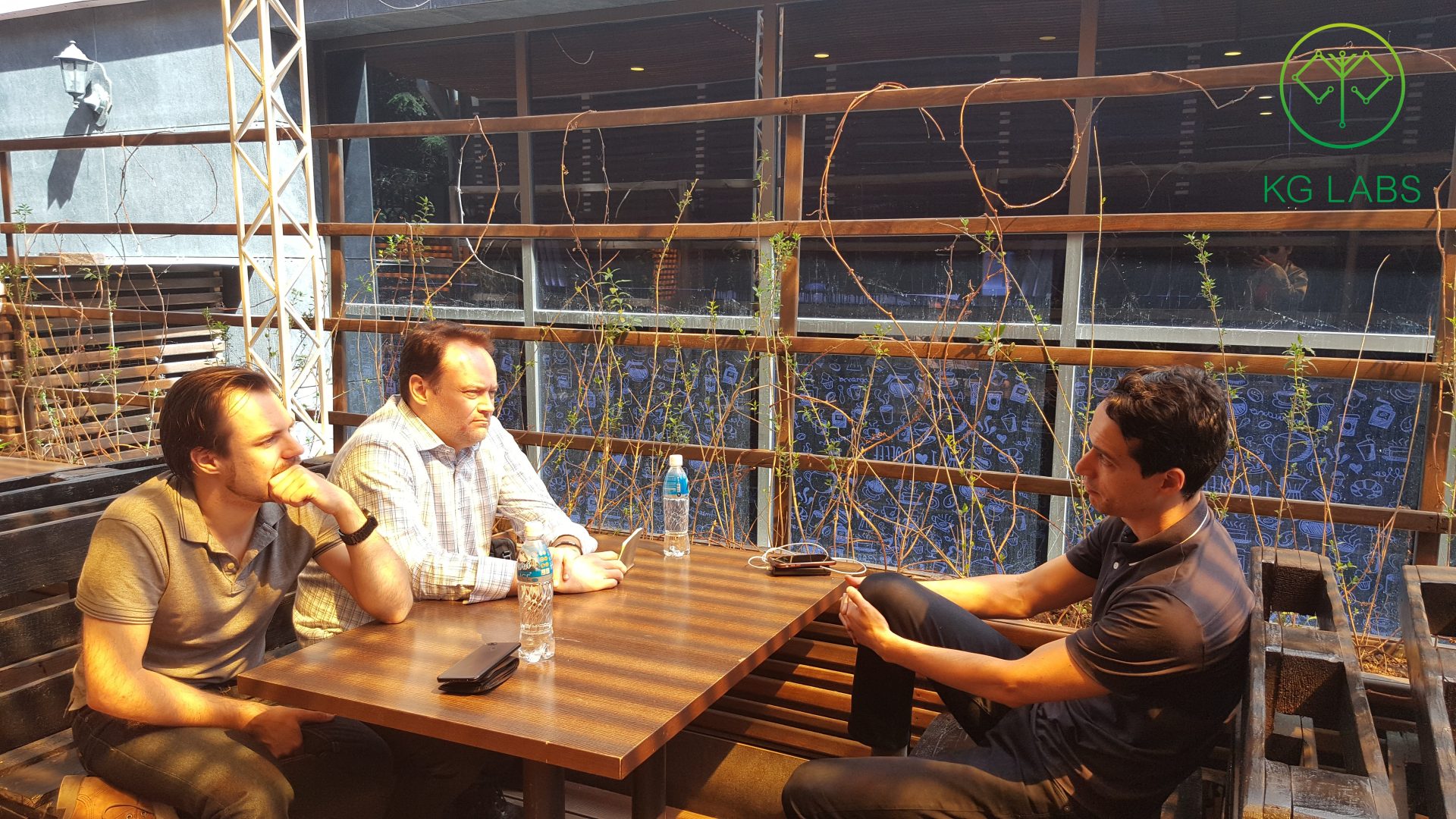 Three individuals engaged in a discussion at an outdoor table with drinks and electronic devices.