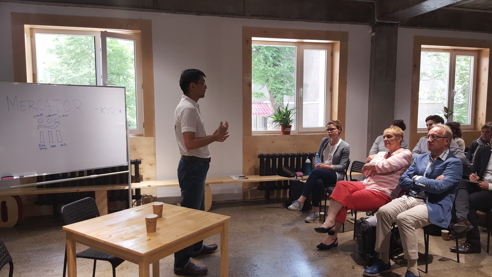 A workshop or training session is taking place in a modern, open-plan office with participants seated and a presenter standing at a whiteboard.
