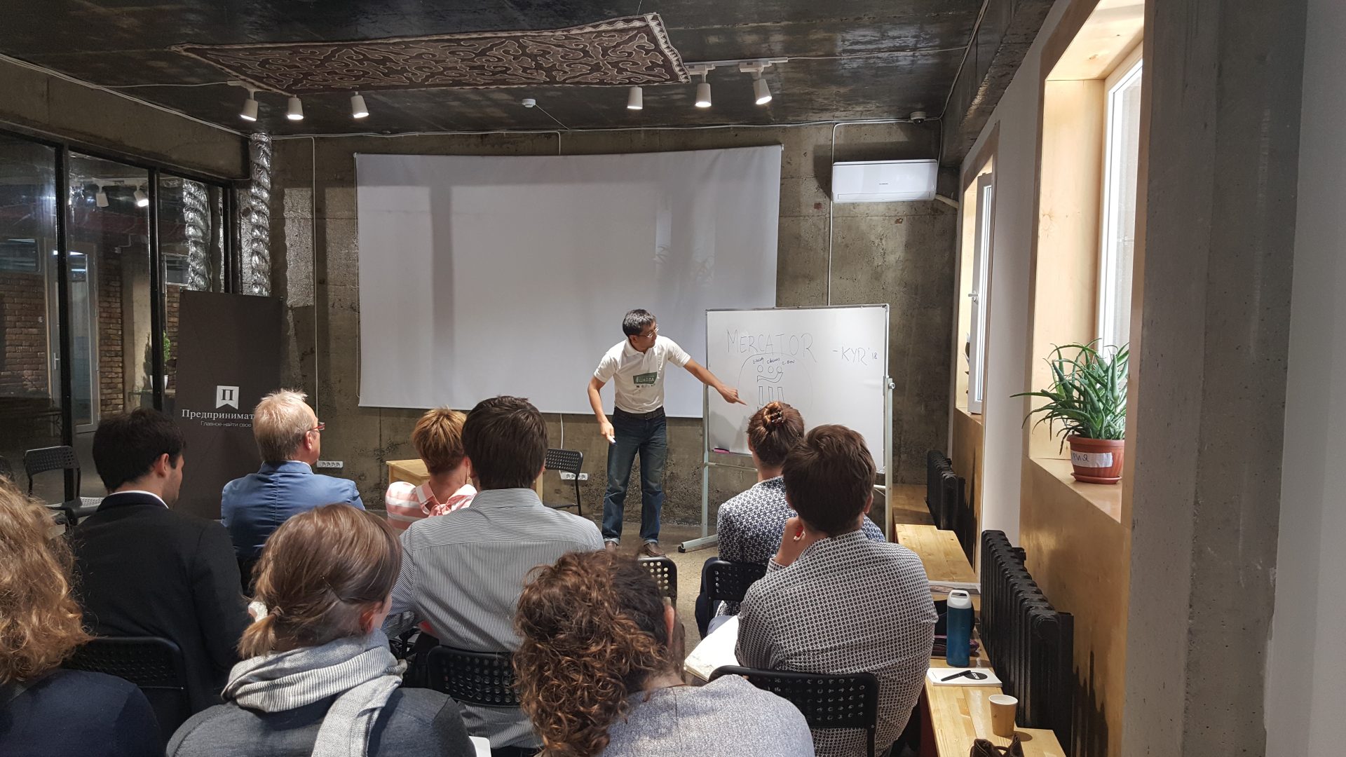 A workshop or training session is taking place in a room with participants seated and a presenter standing at a whiteboard.