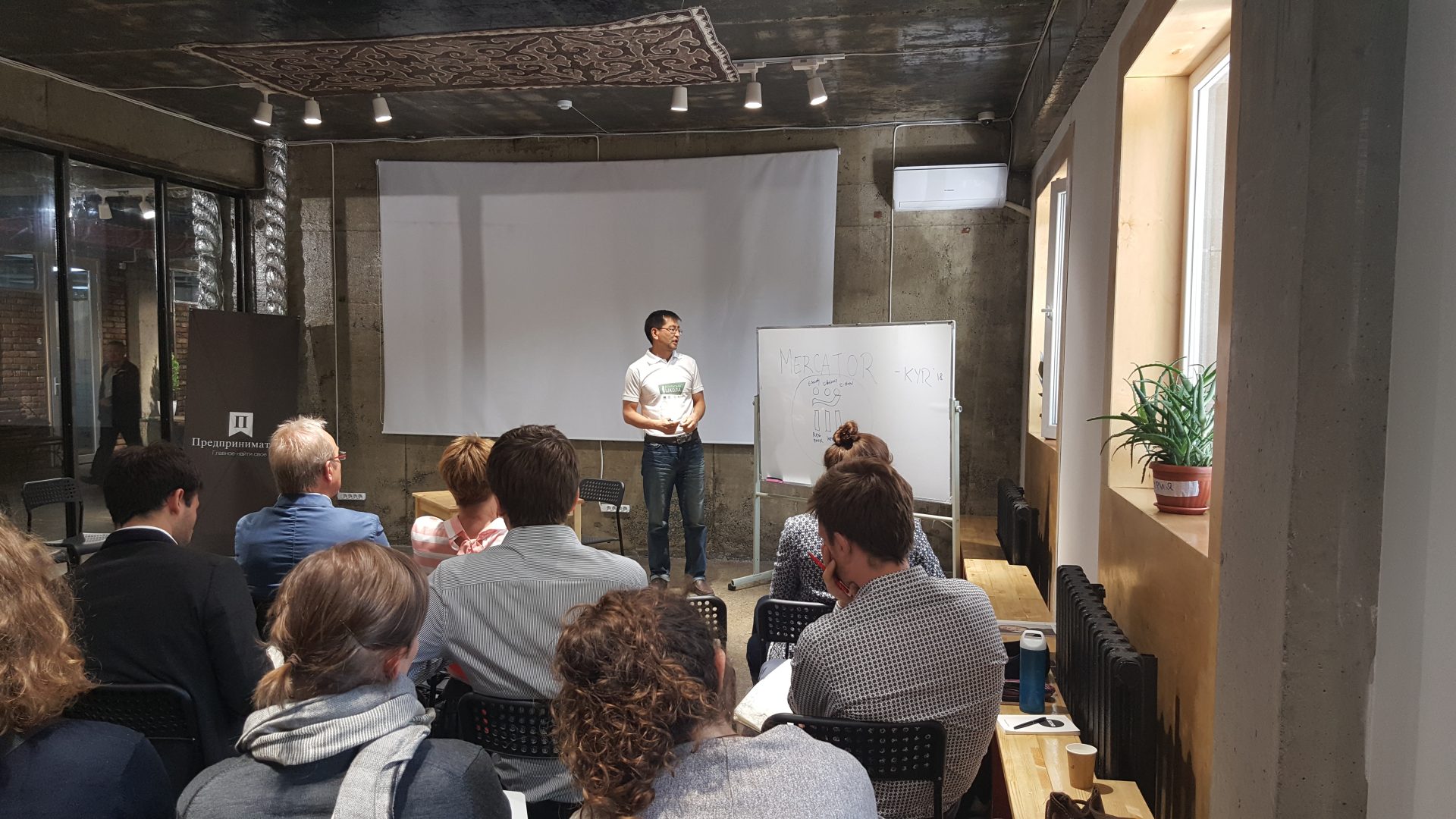 A workshop or training session is taking place in a modern, industrial-style room with participants seated and a presenter standing in front of a whiteboard and projector screen.