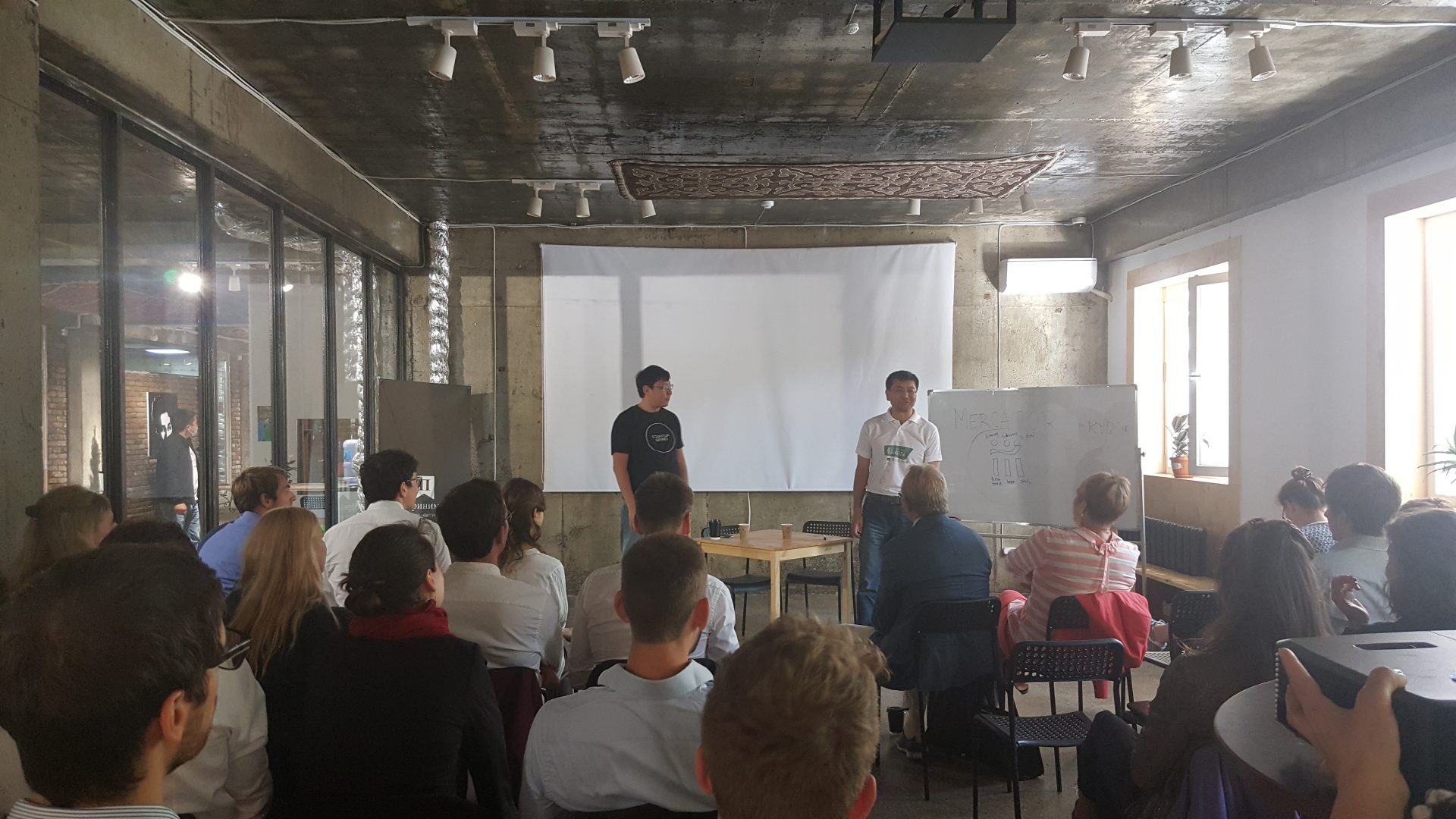 A workshop or training session is taking place in a modern, industrial-style room with participants seated and two presenters standing in front of a whiteboard and projector screen.