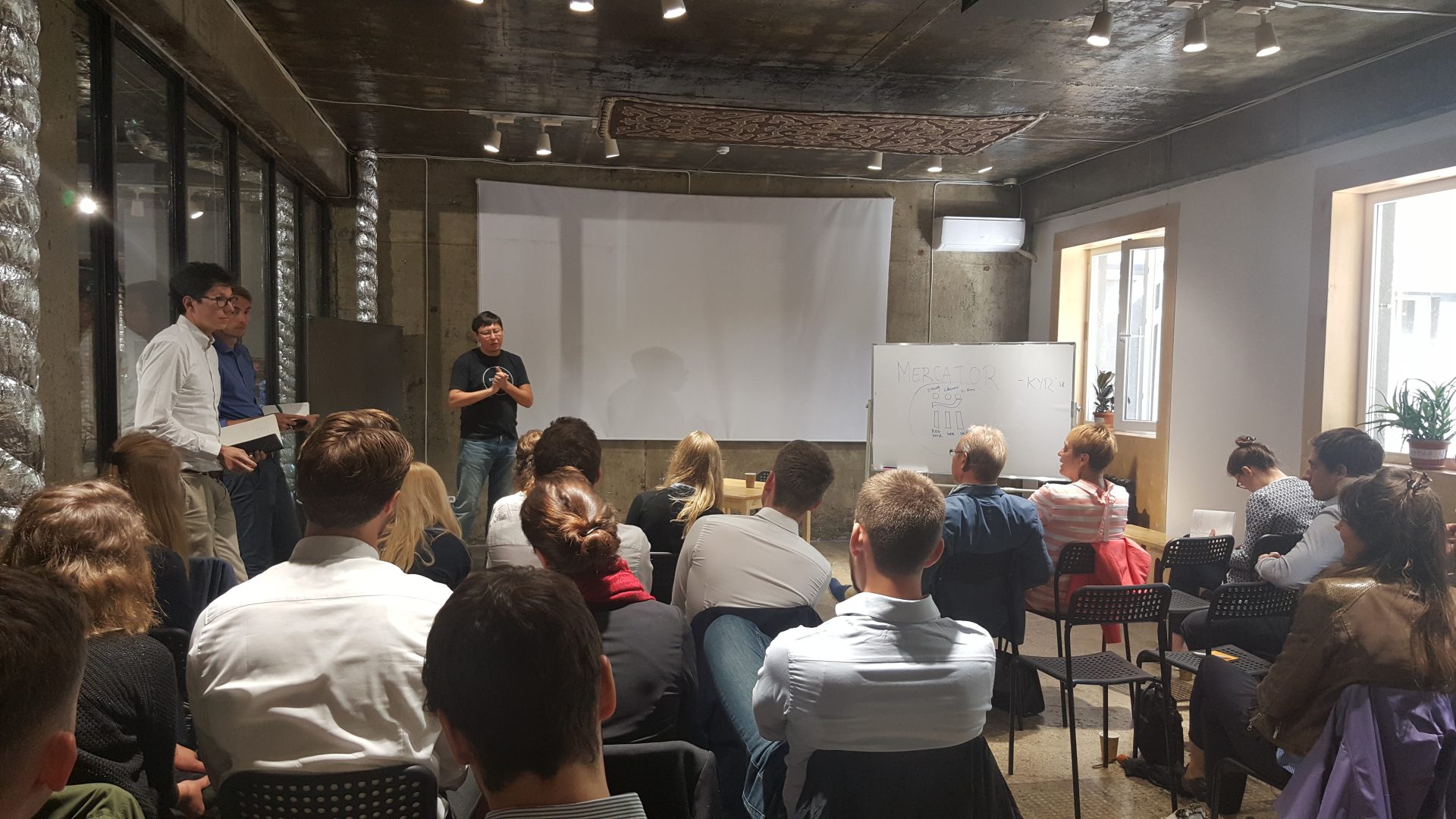 A group of people sitting in a room with a whiteboard and a projector screen, possibly attending a workshop or training session.