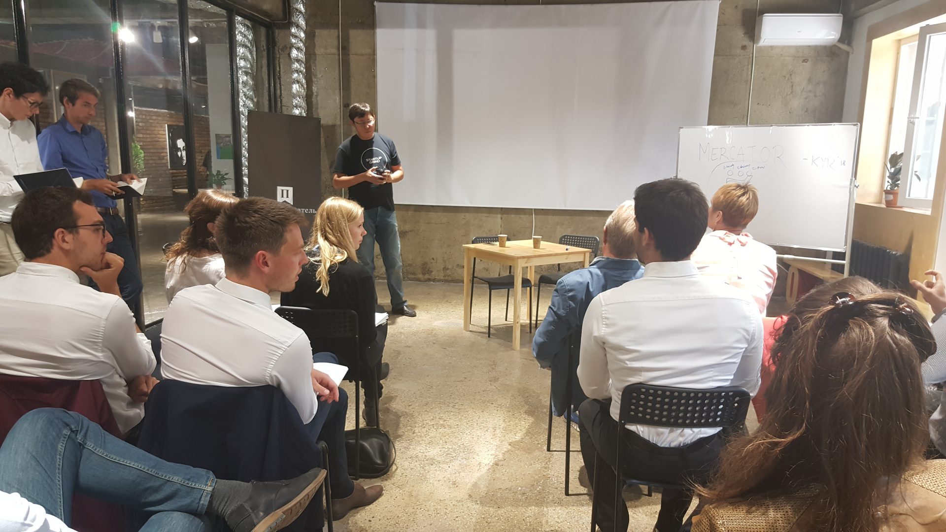 A group of people sitting in a room with a whiteboard and a projector screen, possibly attending a workshop or training session.
