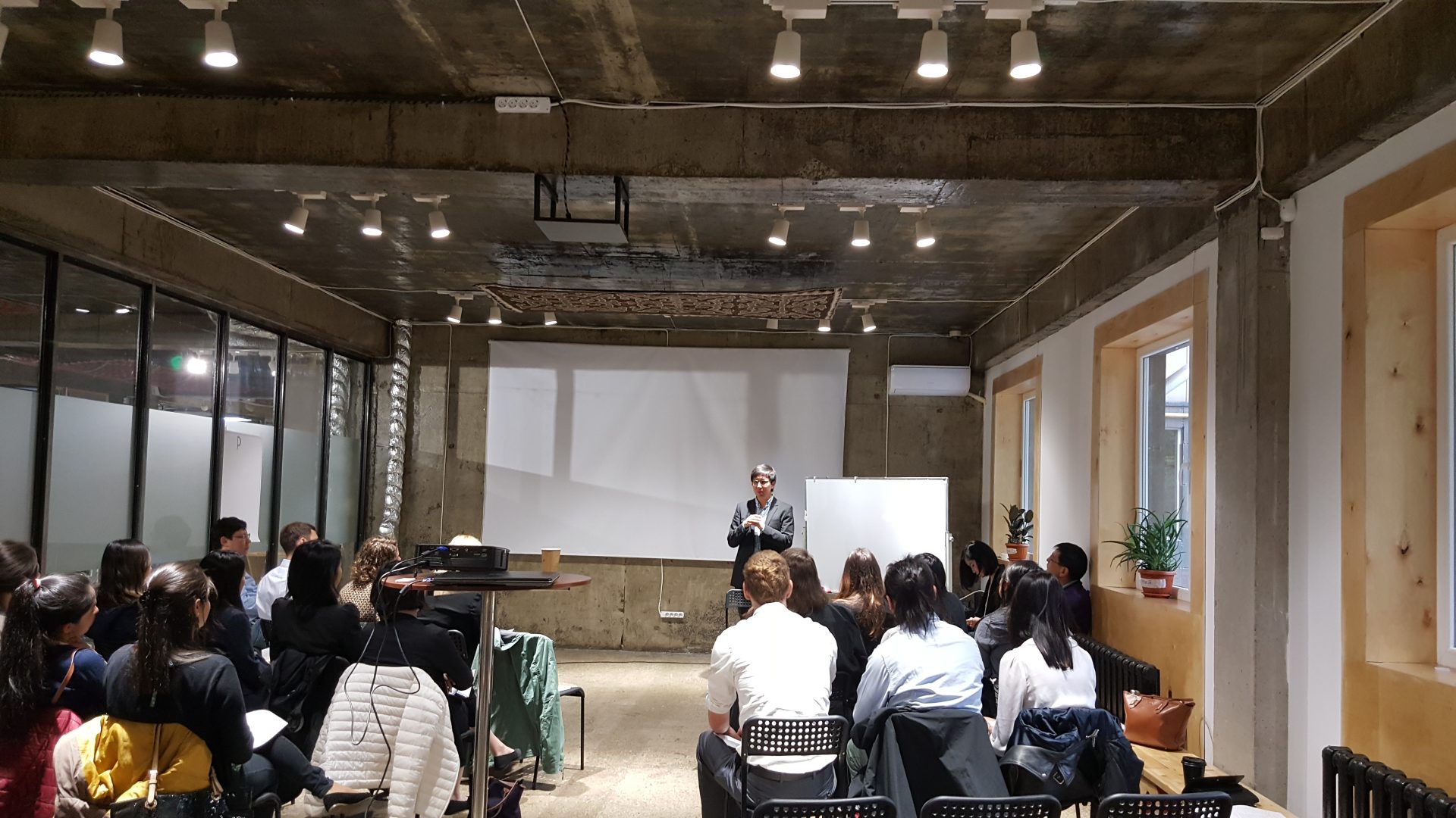 A group of people is seated in a room with a presenter standing in front of a whiteboard and a projector screen.