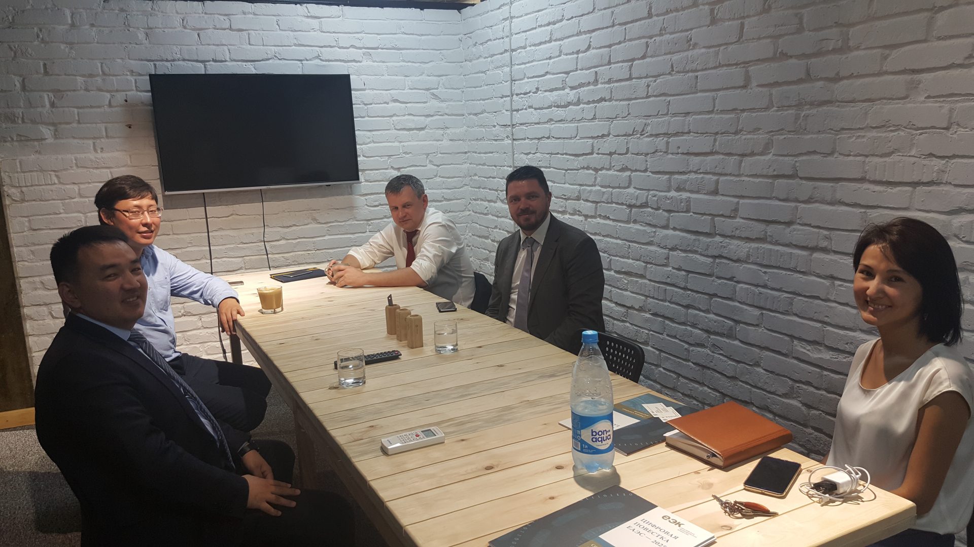Five individuals in a meeting room with a wooden table, white brick walls, and a television screen.