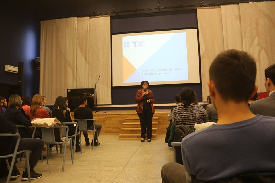 A woman stands on a stage in front of a screen displaying the British Council logo and the words 'Creative Spark: Higher Education Enterprise Programme'.